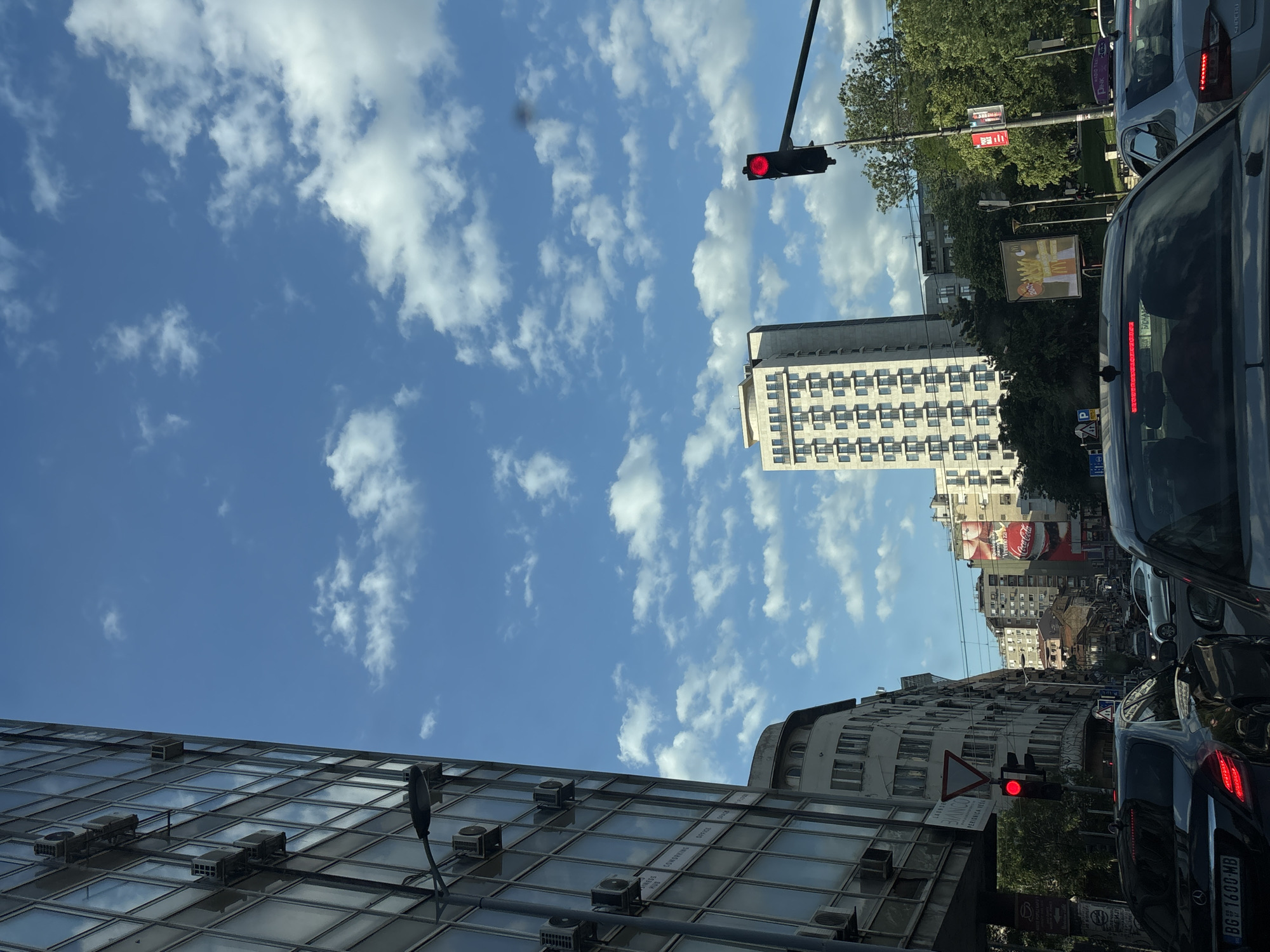 The image depicts a street scene in Belgrade, Serbia, viewed from a car stopped in traffic. The sky is blue with scattered white clouds. Buildings of various heights and architectural styles line the street. A tall, modern building with a grid-like facade and numerous air conditioning units is visible on the left. A taller building with a square, repetitive window pattern stands further down the street. Traffic lights are red, indicating a stop. Cars are visible on both sides of the road. A billboard for McDonald's and a Coca-Cola advertisement on the side of a building are also present. The license plate of a car is visible with the number "BG 1600-MB".