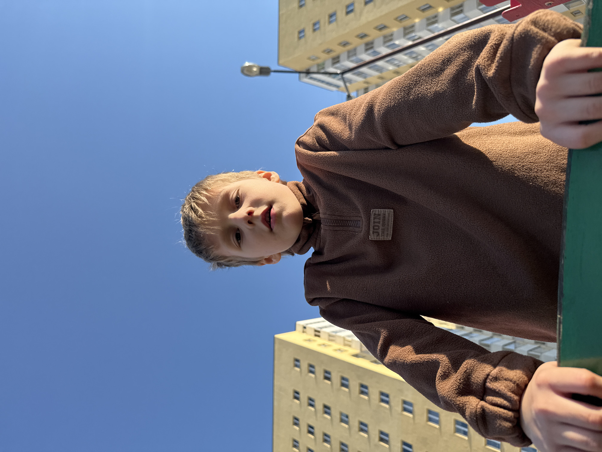 A boy wearing a brown fleece jacket looks down at the camera. He is holding onto a green bar. Behind him are two light yellow apartment buildings. The sky is blue. The picture was taken from a low angle. It is impossible to confirm the location.