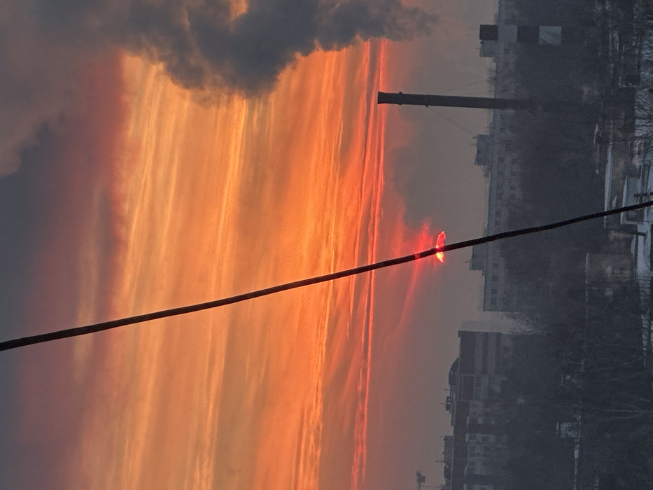 The image depicts an urban-industrial landscape in Kazan, Russia, at sunrise or sunset. A vibrant sky dominates the upper two-thirds, exhibiting intense orange and red hues with horizontal cloud streaks. A dark, thick diagonal cable or pole bisects the frame from the upper-left to the lower-right. Behind this obstruction, a distinct, bright red, half-disk object, consistent with the rising or setting sun, emits a strong glow near the center-left.
The midground features a silhouetted horizon line of buildings and industrial structures. On the left, multi-story buildings and a construction crane are visible. On the right, a tall, dark industrial chimney stands prominently, emitting a large plume of dark grey smoke or steam that rises vertically and disperses into the atmosphere. The ground layer at the bottom appears covered in snow, interspersed with bare trees and bushes, indicating a cold climate. The entire scene is viewed through atmospheric haze. No individuals or specific actions are discernible.