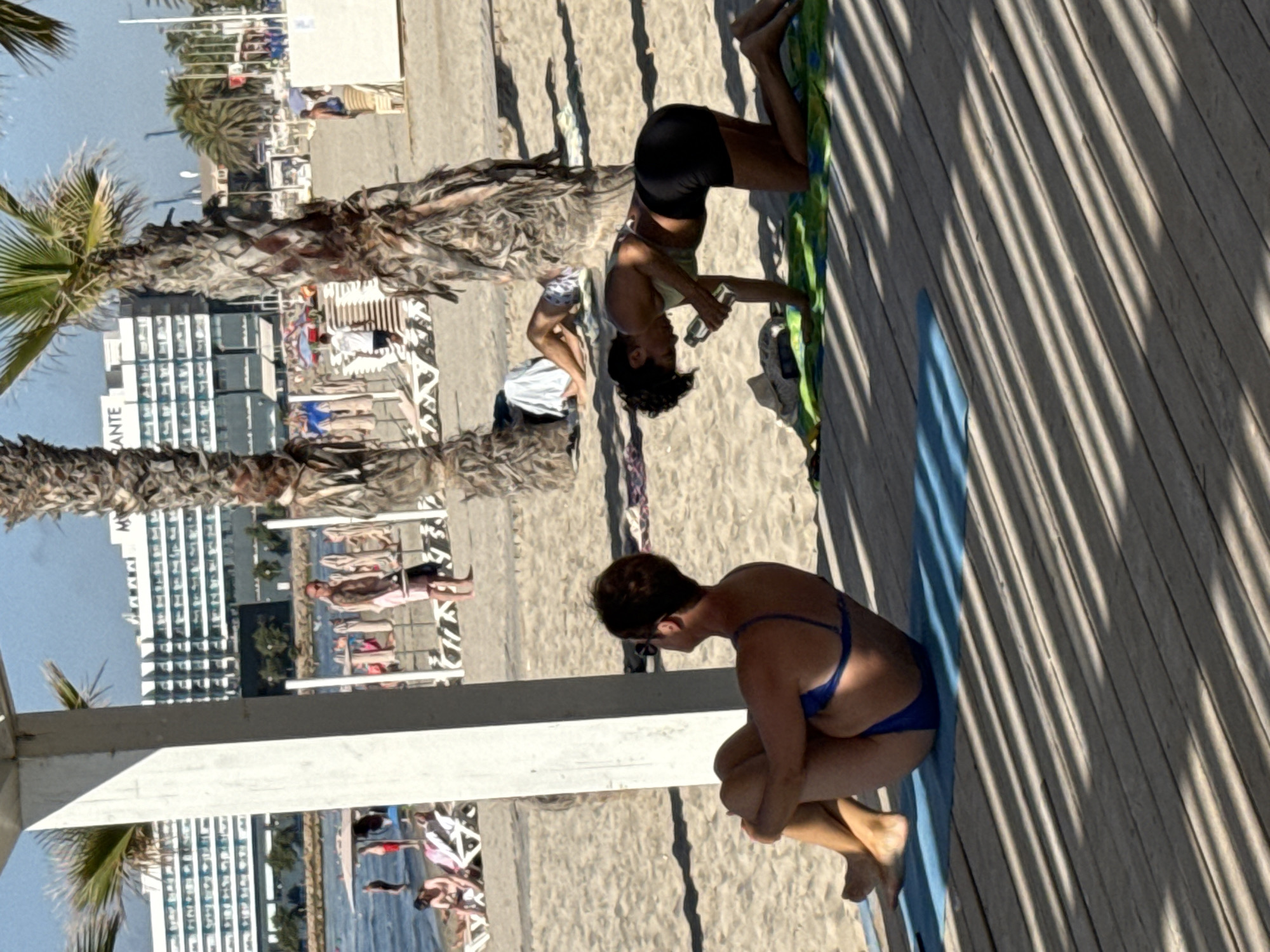 A medium shot captures two women practicing yoga on a wooden deck at a beach resort. In the foreground, a woman in a blue bikini sits on a blue yoga mat, her legs crossed and her arms resting on her knees. To her right, a second woman in a green sports bra and black shorts kneels on a green and blue mat, holding a water bottle. She is in the process of drinking from it. Behind them, palm trees cast striped shadows across the wooden planks. In the background, people are visible on the sandy beach, with lounge chairs and umbrellas. A large, white, multi-story building with many windows is situated behind the beach. The sky is clear and blue.