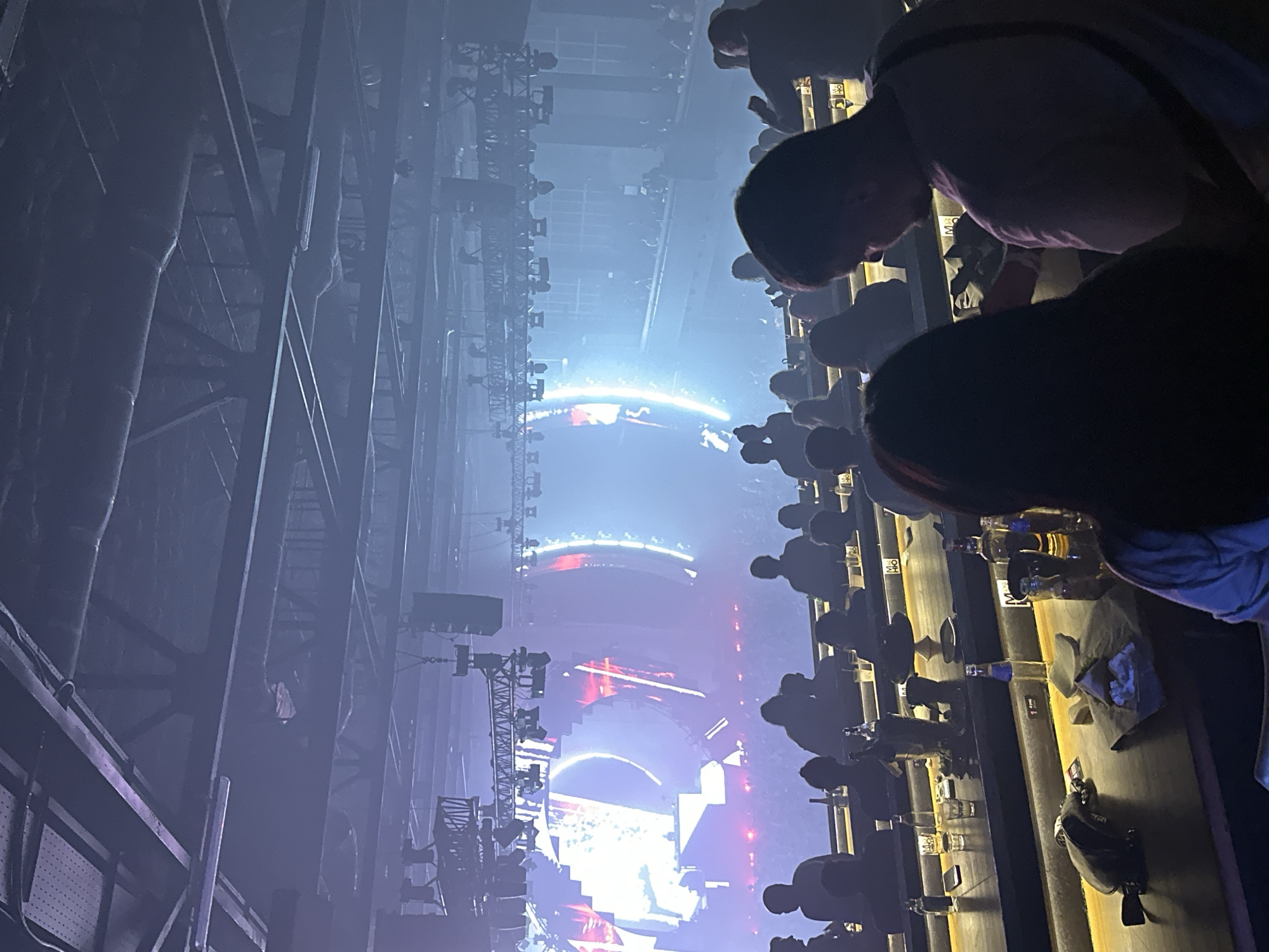 The image captures a bustling indoor concert or club event from an elevated perspective, looking towards a brightly lit stage. The foreground features the heads and shoulders of several spectators, largely silhouetted, some leaning over a bar or counter. On this counter, various bottles, including some containing amber liquid and one with a blue label, along with glasses and a small black bag, are visible. A small white sign with what appears to be "HO" or "OH" is also on the counter.
The venue itself is dark and atmospheric, possibly hazy from stage effects, with structural beams and scaffolding prominent on the left side, suggesting an industrial aesthetic or temporary event setup. Rows of additional spectators fill the mid-ground, appearing mostly as dark figures against the brighter backdrop.
The focal point is the distant stage, dominated by massive, glowing circular and semi-circular LED screens or lighting fixtures emitting brilliant white, blue, and red light. These light structures create dynamic patterns, hinting at a vibrant performance underway. Truss systems and professional stage lighting rigs are also visible around the stage area. The overall impression is of an energetic evening event, likely a music performance, in a large, modern entertainment venue in Khimki, Russia.