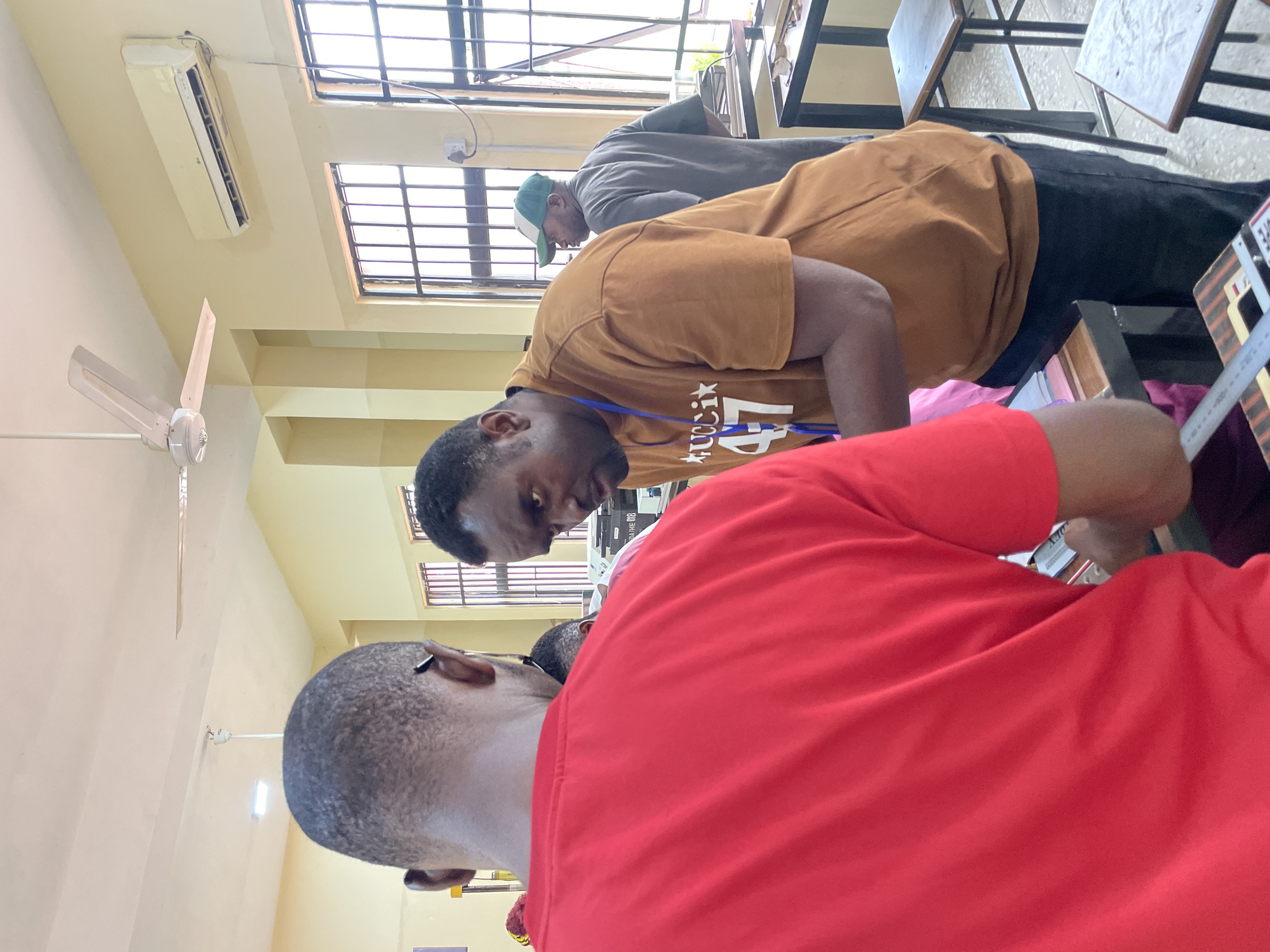 Three men are indoors, possibly in a classroom in Afaka, Nigeria. One man is wearing a red shirt and is closest to the camera, with only the back of his head and upper back visible. Another man is wearing a tan t-shirt with writing on it and a blue lanyard around his neck, leaning forward, possibly looking at a document or object on a table. The third man is wearing a gray shirt and a green and white baseball cap, also leaning forward and appearing to look at something on the table. The room has yellow walls, windows with bars, a ceiling fan, and an air conditioning unit. There are tables and chairs in the background.