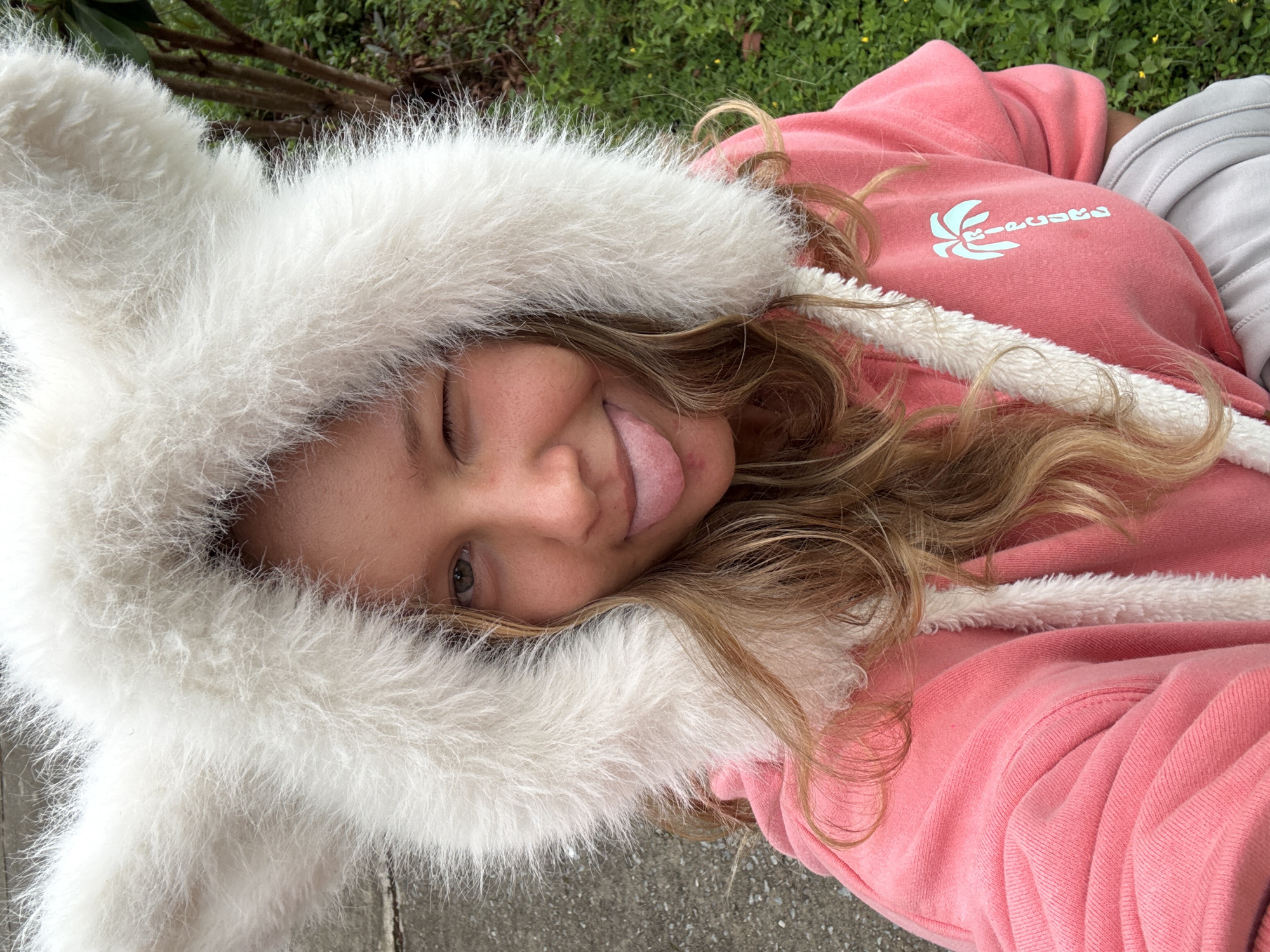 A young woman with blonde hair is wearing a pink hooded sweatshirt with white faux fur trim. The hood is up and has large, fluffy white ears, resembling those of a rabbit. She is winking and sticking her tongue out at the camera. She is wearing a grey pair of sweatpants. The background shows a paved surface on the left and lush green vegetation, including trees and grass, on the right. The image appears to be taken outdoors, likely in a residential or park area. The location is unspecified.