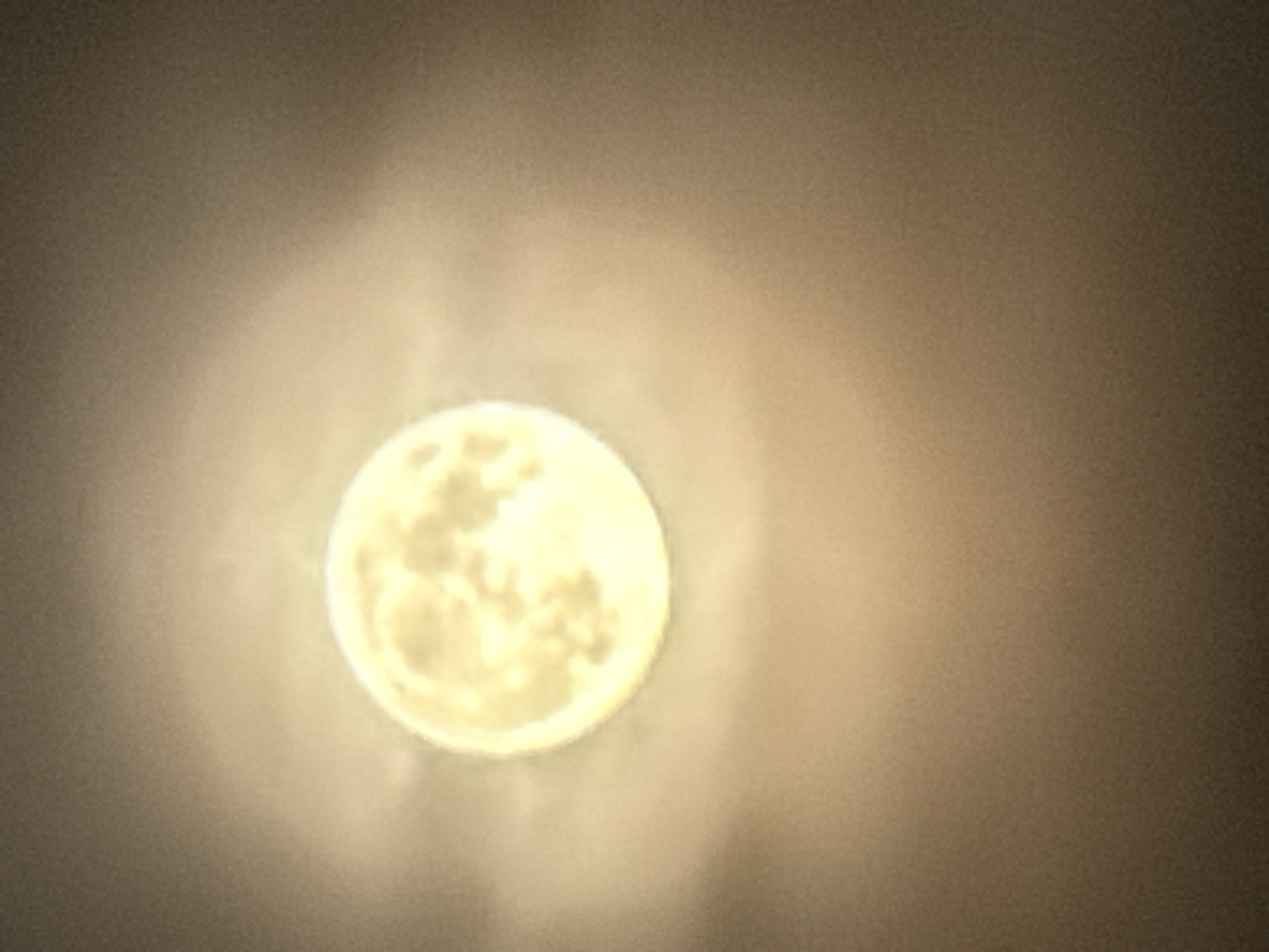 The image shows a full moon with a hazy glow around it. The moon is a bright, yellowish-white sphere with some visible craters or surface features. It is set against a dark, out-of-focus background that appears to be a night sky with some atmospheric haze or light pollution. The overall impression is that of a celestial body observed from Earth. The image does not depict any specific location in the city of Kamianka-Buzka, Ukraine, nor does it show any people, objects, actions, or interactions.