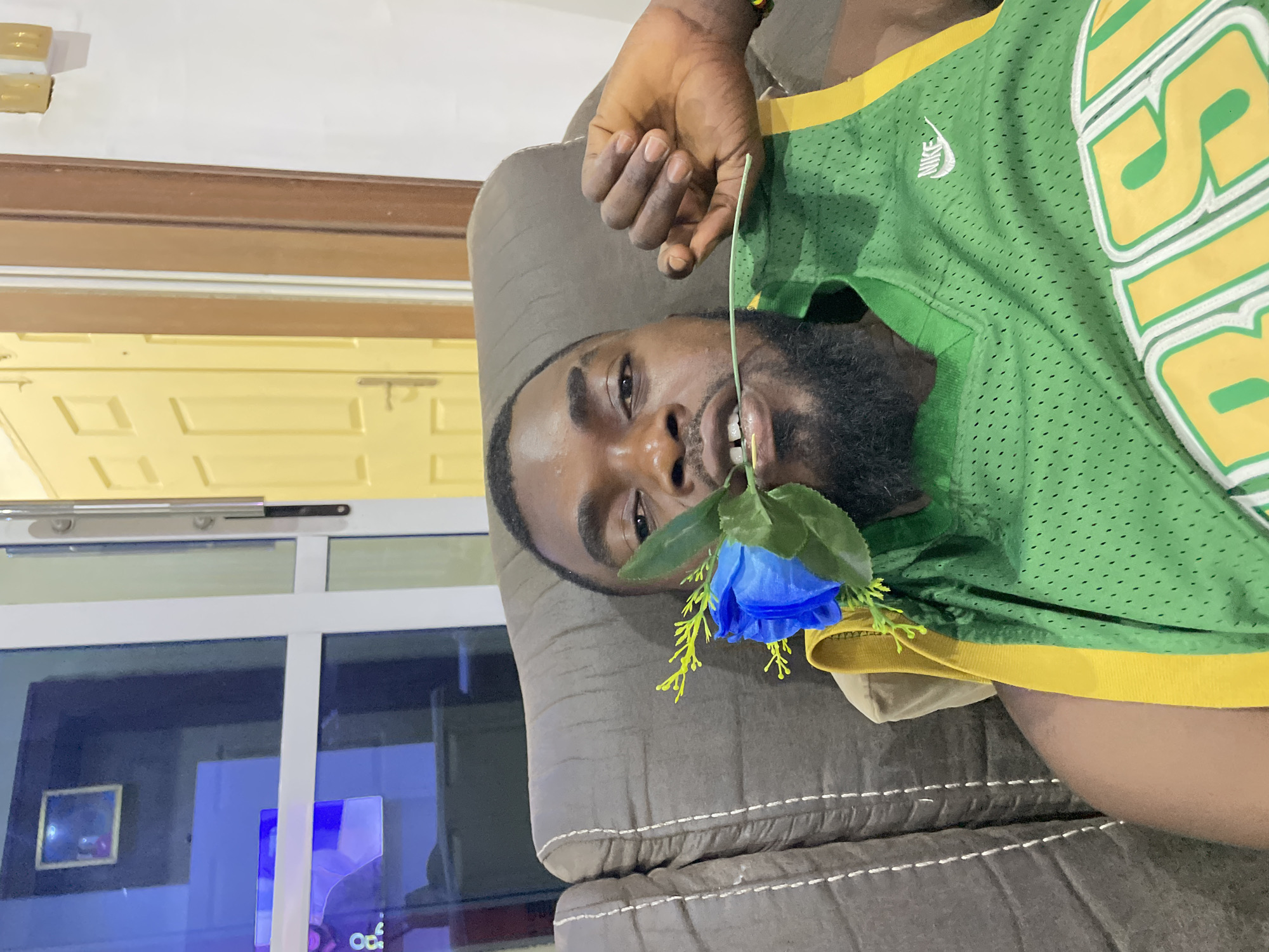 A man with dark skin, a beard, and short dark hair is shown lying on his side on a dark gray upholstered sofa. He is looking directly at the camera with a slight smile. In his mouth, he holds a vibrant blue artificial rose with green plastic leaves and yellow accents.
He is wearing a green mesh sports jersey with yellow trim on the collar and sleeves. A small white Nike logo is visible on the left side of the chest, and large yellow letters outlined in white and green, partially spelling "BIG" or a similar word, are emblazoned on the right side. A multi-colored bracelet is visible on his right wrist.
The background of the indoor setting includes a pale yellow wall or door with raised panels, and a brown wooden door frame or trim. A white ceiling with a light fixture is seen above. To the lower left, a window or glass door reflects a dark interior space, showing a framed picture and an area emitting a blue light, possibly from an electronic screen. The lighting appears to be artificial, suggesting it might be evening or nighttime. The man's expression is relaxed and playful.