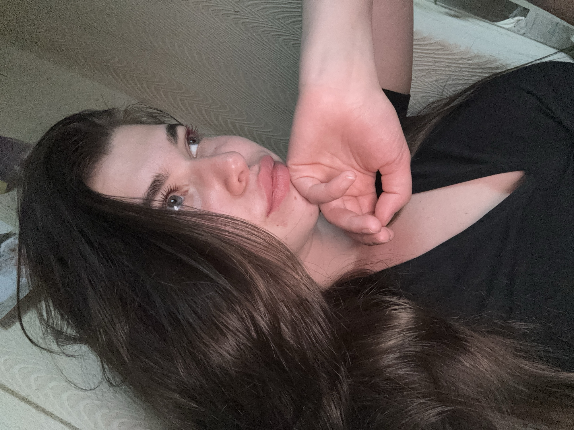 A young woman with long, dark brown hair is lying down on a surface with a beige patterned texture. She is wearing a black shirt. Her left hand is positioned near her face. The image is taken from a slightly elevated, tilted angle. There is no visible evidence to place this image in Мещёры, Russia.
