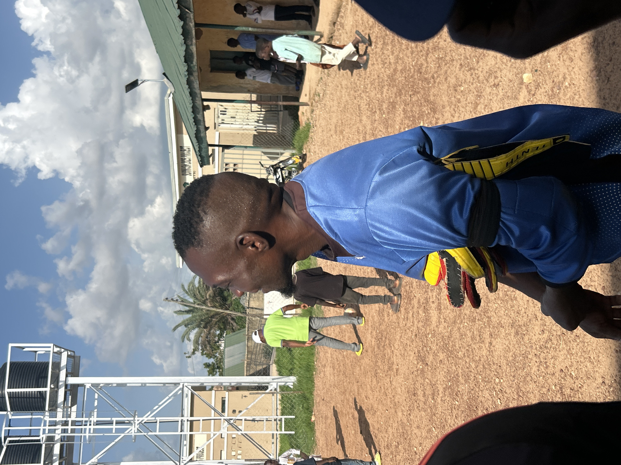 The image captures an outdoor scene in the city of Jos, Nigeria, under a bright sky with scattered white clouds. The ground is composed of reddish-brown earth.
In the immediate foreground, a dark-skinned man is visible in profile, facing left, appearing to smile or laugh. He has a short, faded haircut and is wearing a blue long-sleeved athletic shirt, which has a dotted pattern on its lower section and a black band on his left upper arm. He holds multiple items on his left arm, appearing to be athletic gloves or pads, which are colored red, yellow, and black; one yellow item displays the word "TENTH". Portions of two other individuals, identified by their shoulders, are present in the extreme left and right foreground.
In the midground, a man in a bright green t-shirt, gray pants, and yellow shoes stands with his back largely to the viewer, facing right. He is near a dark-skinned boy in a black t-shirt and dark pants, who is bent over a soccer ball on the ground. A metal mesh fence separates this area from a patch of grass and a yellow and black motorcycle parked behind it.
The background features several structures and vegetation. To the left, a tall, white metal water tower supports two black water tanks. Behind the water tower and to its right, light-colored buildings are visible; one building has a sign that partially reads "PROJECT". To the right, a single-story building with light-colored walls and a green corrugated metal roof with an overhang is present. A solar-powered light fixture is mounted on this roof. Several individuals are standing or walking near the entrance of this building, including one person in a light blue shirt and light-colored pants moving towards the left. Palm trees and other green foliage are distributed throughout the background. The scene is illuminated by strong sunlight, casting visible shadows on the ground.