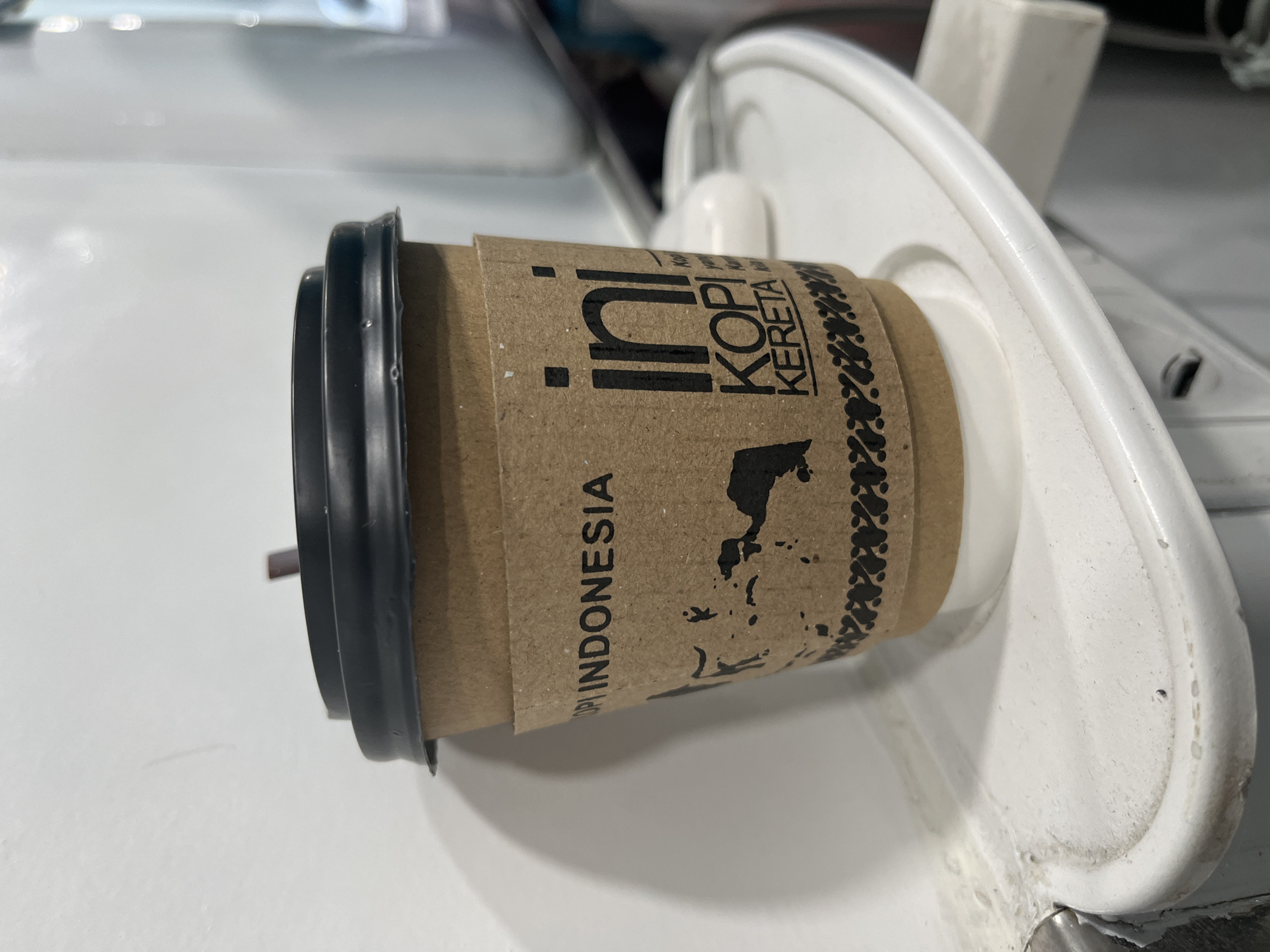 A close-up, eye-level shot shows a paper coffee cup with a black lid and a brown sleeve. The sleeve has black text and a silhouette of Indonesia. The text reads "KOPI INDONESIA" and "ini KOPI KERETA." The cup is sitting on a white tray, which is mounted on a white surface that appears to be the interior of a vehicle, possibly a train, given the text on the cup. The background is blurred and white. The location is not specified in the image or implied.