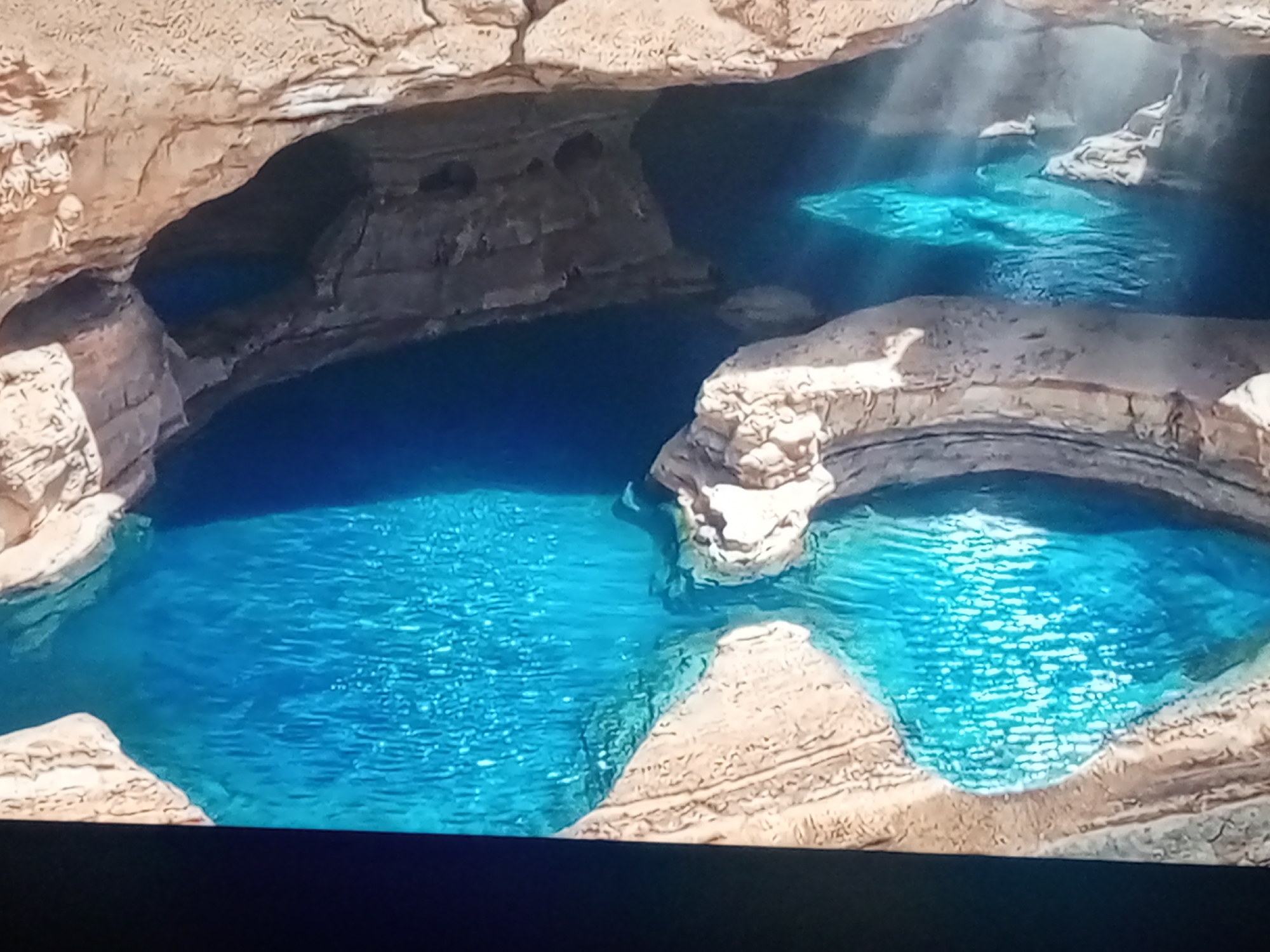 The image shows a series of bright blue pools of water nestled within a cavernous landscape. The rock formations surrounding the pools are a light brown and appear to be carved by water erosion, with visible layers and rough textures. Sunlight streams down into some of the pools, creating illuminated patches and reflections on the water's surface. Some of the pools are interconnected, with water flowing between them. The overall impression is one of natural beauty and tranquility. The location is described as Unknown, Unknown.