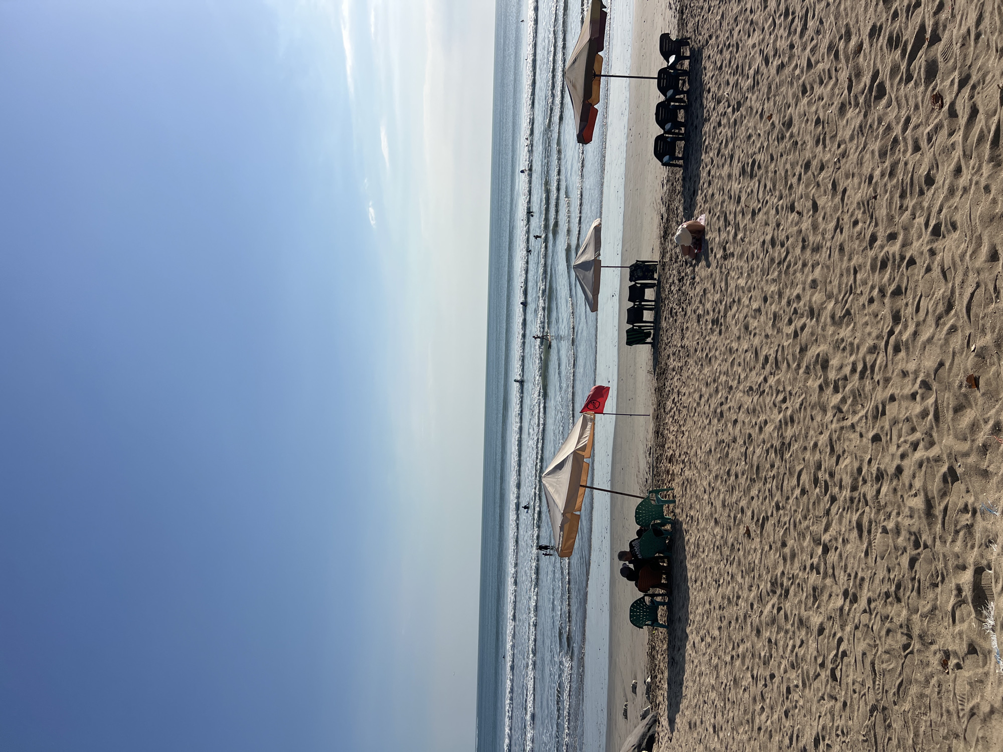The image depicts a serene beach scene in Kuta, Indonesia. The foreground is dominated by a sandy beach with visible footprints and some scattered dry leaves. In the mid-ground, several beach umbrellas provide shade, with lounge chairs arranged underneath them. Two umbrellas are positioned together, with a group of people seated in chairs under one, facing the ocean. A red flag is planted near this group. Further to the right, another umbrella stands over a set of chairs, and a solitary individual is seen kneeling on the sand beneath it. The background features the ocean with gentle waves, and numerous surfers are scattered across the water. The sky is a clear, bright blue, indicative of a sunny day.