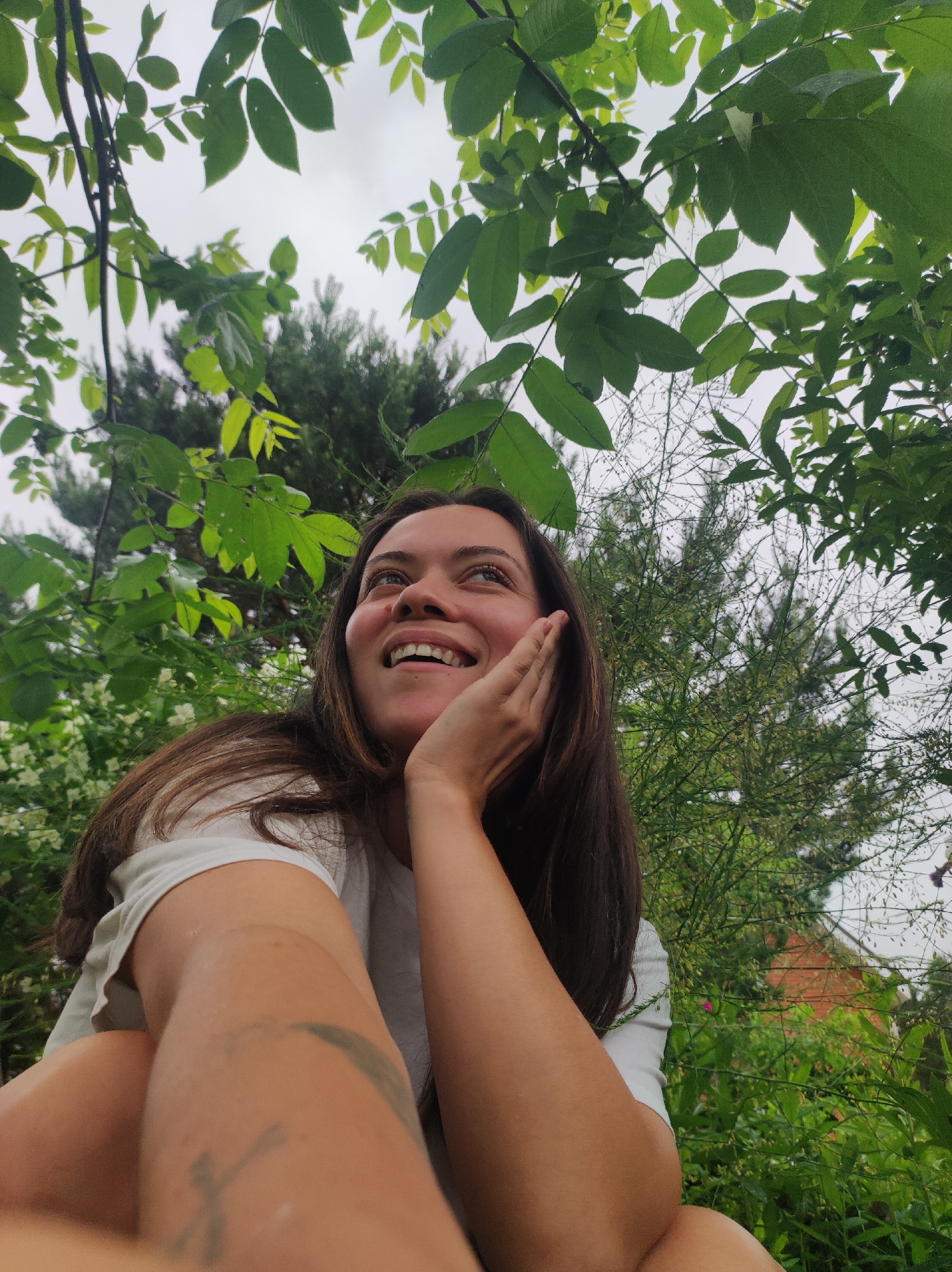 The image features a young woman captured in an outdoor setting in Фоки, Russia, looking upwards with a joyful and wonder-filled expression. She has long, dark brown hair and is wearing a white t-shirt. A wide, toothy smile is evident on her face, and her right hand gently rests against her cheek. Her left arm and part of her leg are visible in the foreground, with a faint, dark, abstract tattoo on her left forearm.
The photograph is taken from a low-angle perspective, looking up through a dense canopy of vibrant green leaves and branches, which frame the upper part of the sky. The sky appears overcast, suggesting diffused, soft light. Beyond the immediate leafy foreground, various other green bushes and trees, including some with small white blossoms, form a lush natural backdrop. A blurry, reddish-orange structure, possibly a building or house, can be seen in the distant background on the right, nestled amidst the foliage. The scene conveys a sense of peace and connection with nature on an overcast day. No discernible text is visible on clothing or in the surroundings, other than the tattoo.