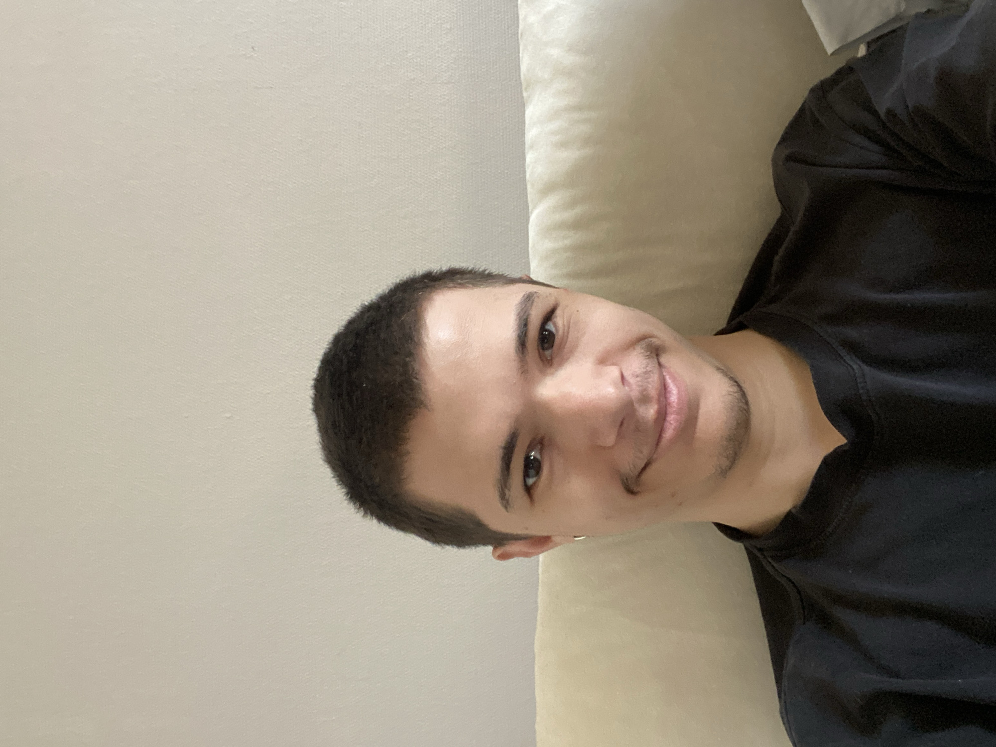 A young man is featured in a close-up, selfie-style portrait, likely taken while he is reclining or lying down indoors. The image is tilted approximately 90 degrees to the right, showing his head and upper torso. He has short, dark, buzz-cut hair, dark eyes, and subtle stubble forming a small goatee. A gentle, relaxed smile is visible on his lips, conveying a sense of contentment or ease.
He is wearing a plain black t-shirt. The background consists of a light, neutral-colored wall or plain surface to his left and a plush, textured, light-colored pillow or cushion to his right, suggesting he is on a couch or bed. The lighting is soft and even, characteristic of an indoor setting, making it difficult to determine the exact time of day. There is no visible text in the image. The scene depicts a casual, relaxed moment, located in Strasbourg, France.