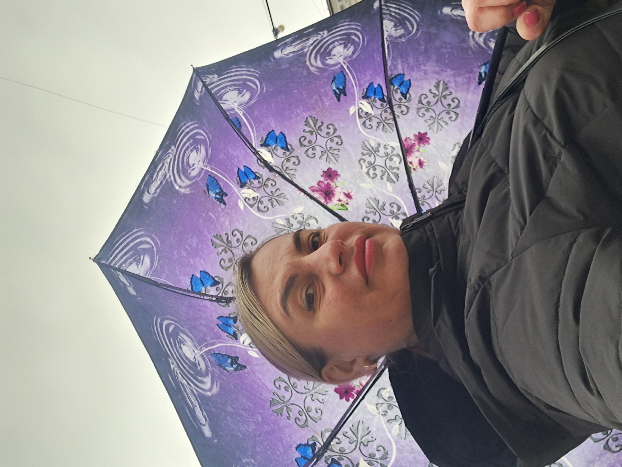 A woman stands under a purple and blue patterned umbrella. The image is taken from a low angle, looking up at the woman and the umbrella. The woman has light skin, blonde hair, and is wearing a black quilted jacket. The background appears to be a cloudy sky or a light-colored building. There is no explicit information to confirm or deny if the picture was taken in the city of Elektrostal, Russia.