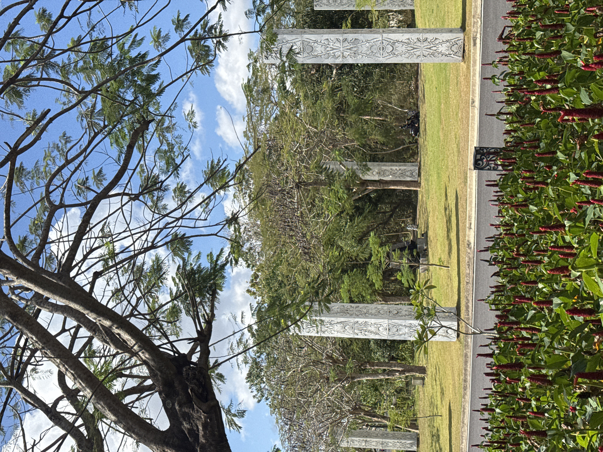 The image shows a street in Kuta Selatan, Indonesia, lined with a vibrant flower bed of red ginger flowers in the foreground. Beyond the flowers, a road separates the viewer from a grassy area featuring several tall, ornate white pillars with intricate carvings. In the background, lush green trees and foliage create a dense natural backdrop. The sky above is a clear blue with scattered white clouds. The scene appears peaceful and serene, with a light motorcycle parked on the grass near one of the pillars, suggesting a public or park-like area.