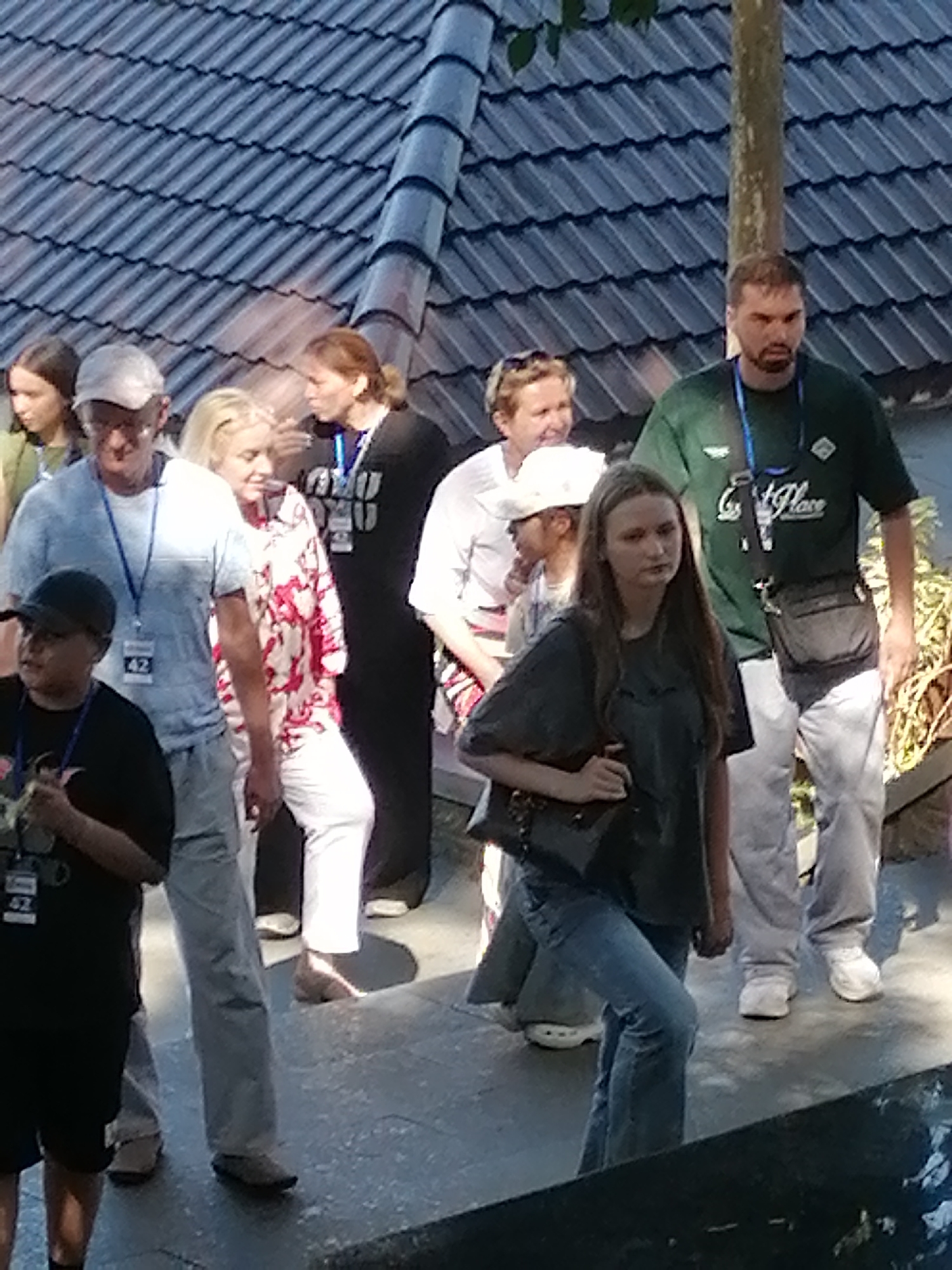 The image depicts a group of people walking on a tiled walkway with a dark, segmented roofline overhead. A tree trunk is visible on the right side of the frame. Several individuals are captured in motion, with varying clothing styles and expressions. In the foreground on the left, a young person wearing a black t-shirt and shorts stands looking to the left. To their right, a man in a light blue shirt and light-colored pants, wearing a hat, is walking and looking forward. Next to him, a woman in a white and red floral pattern shirt and white pants is walking and looking to her right. Further back, a man in a green t-shirt and grey pants is walking and looking forward. A young woman in a dark grey t-shirt and blue jeans is also walking and looking to her left, carrying a black bag. The overall scene appears to be outdoors during the daytime. There is no discernible visual information within the image that can be used to determine the specific location in Da Lat, Vietnam.