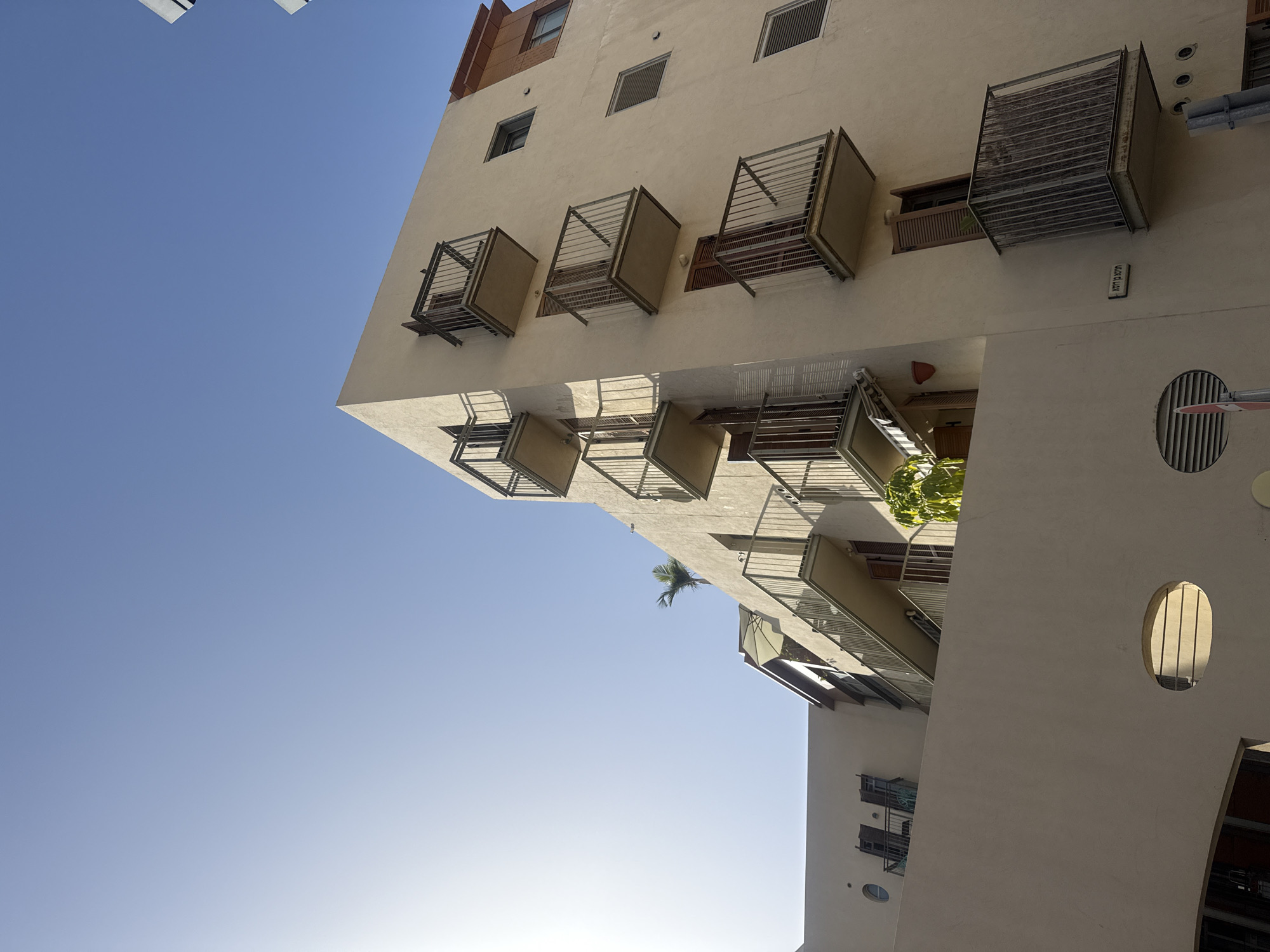 The image depicts a low-angle view of a building exterior under a clear blue sky. The building appears to be located in Tel Aviv, Israel, as indicated by a street sign in Hebrew, which reads "Yisha'ayahu Street". The building is of a light beige color and features several balconies with metal railings protruding from its facade. It also has a series of windows and distinct oval-shaped openings along its side. A single palm tree can be seen in the middle of the building. The lighting suggests it is daytime, and the sun is positioned behind the building, casting shadows on the structure.