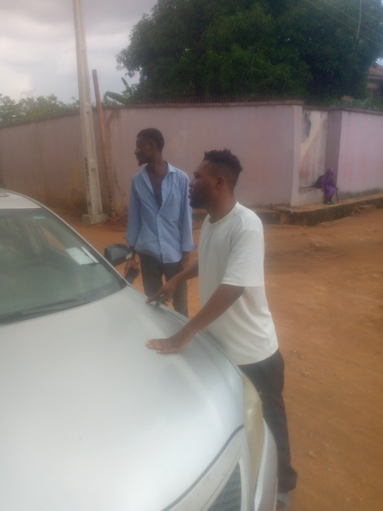 The image shows two men standing near a silver-colored car on a street in an urban environment. The man in the foreground is wearing a white t-shirt and dark pants, with his hand resting on the hood of the car. Behind him, another man is wearing an unbuttoned light blue shirt and dark pants. A concrete wall runs along the back of the scene, with a person sitting on it in the distance. A utility pole stands near the wall, and a tree is visible over the wall. The ground appears to be dirt or reddish soil.