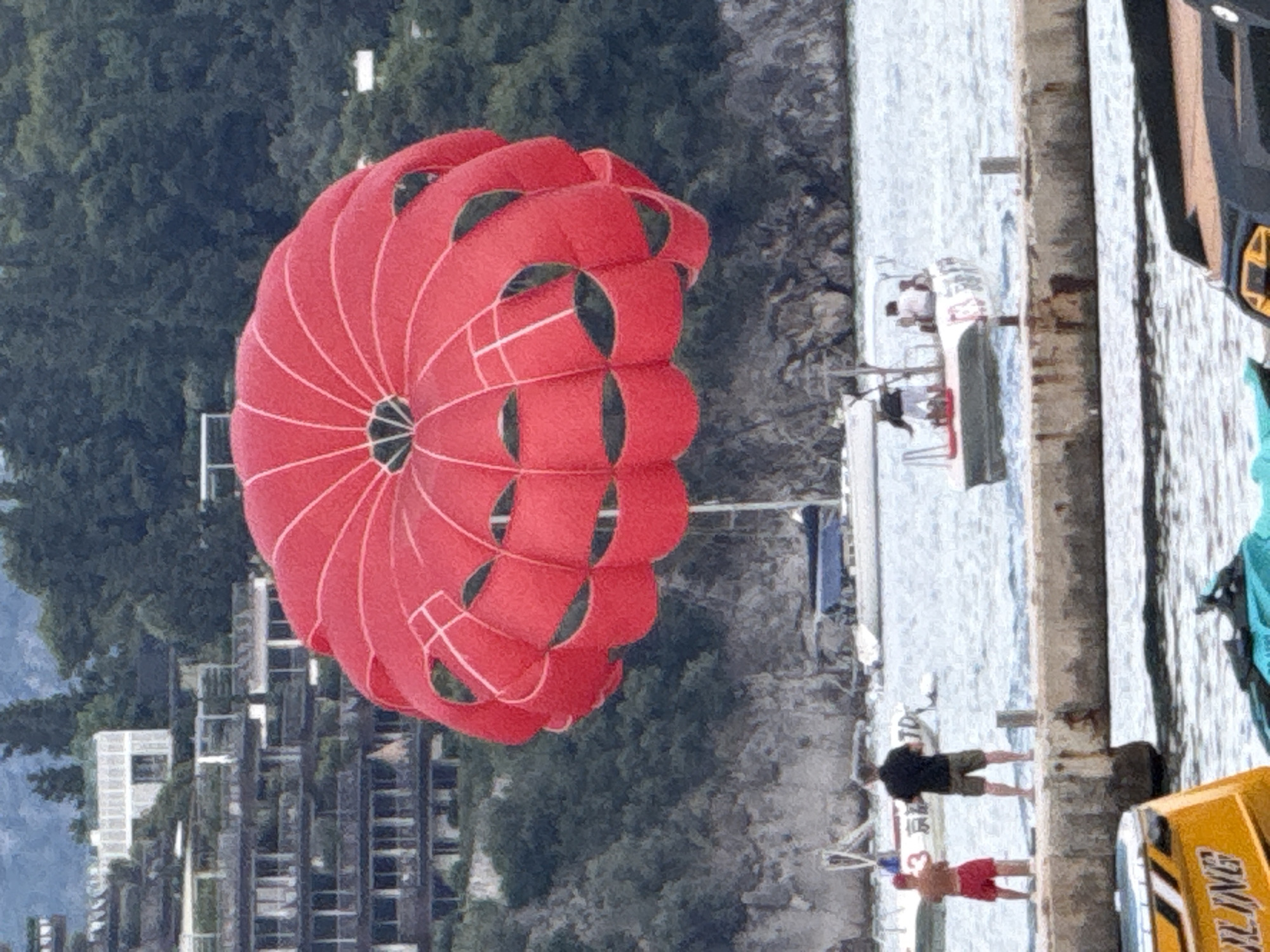 In Budva, Montenegro, a bright red parachute, likely used for parasailing, dominates the foreground of the image, suspended in the air against a backdrop of lush green hills and buildings. Below, a body of water is dotted with boats, including a white sailboat in the middle distance and a smaller motorboat with people on board. A concrete pier runs along the bottom edge of the image, with two men standing on it. One man, wearing red shorts, faces away from the camera, while the other, in dark clothing and khaki shorts, also faces away. A yellow boat with "LING" visible on its side is partially in view at the bottom left, and a blue jet ski is visible at the bottom right. The architecture on the hillside suggests a resort or residential area overlooking the water. The overall scene depicts a leisurely day at the coast.