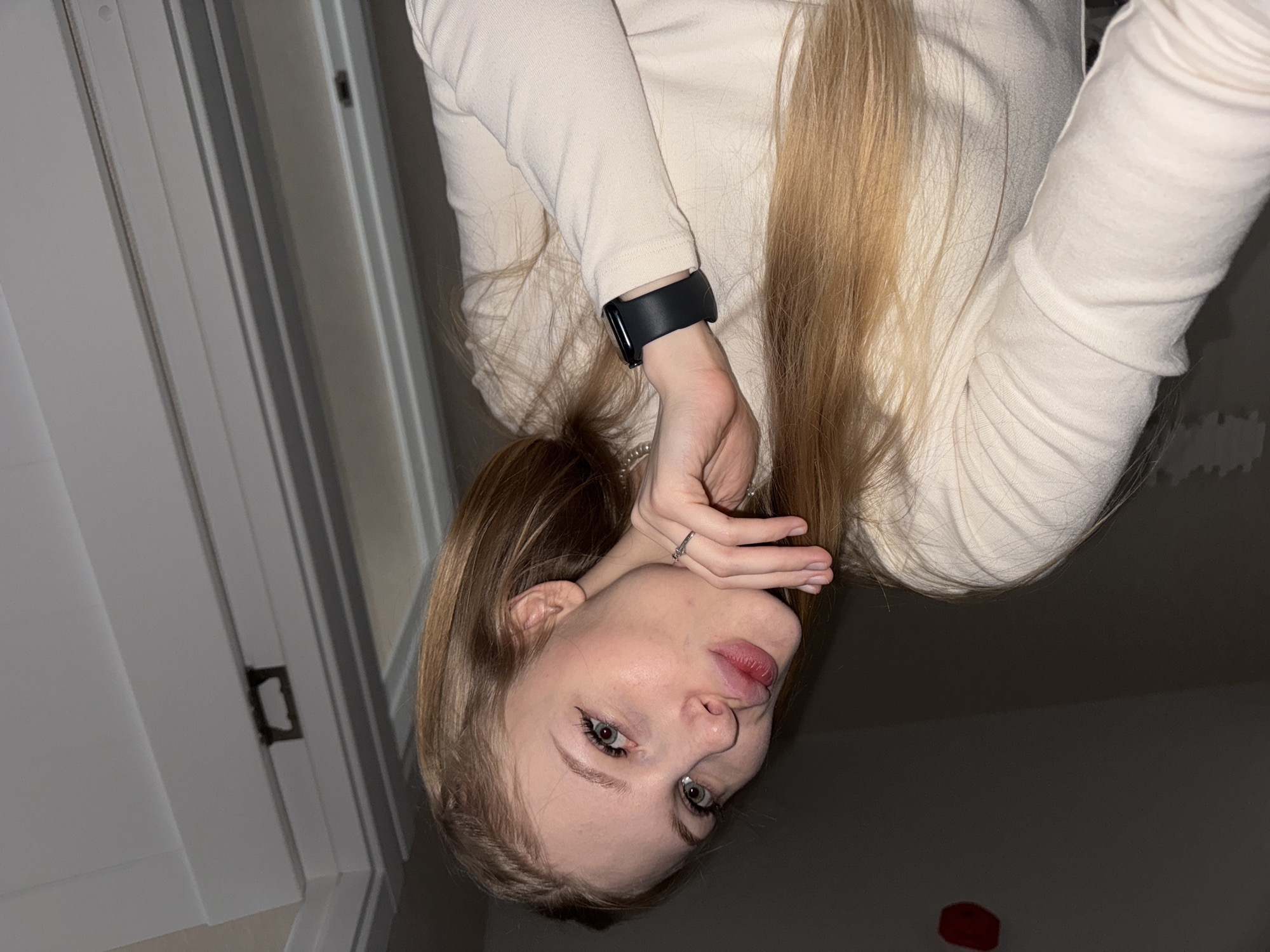 A young Caucasian female with long blonde hair and green eyes is pictured in a close-up, forward-facing shot. She is wearing a light-colored, long-sleeved shirt and a black watch on her left wrist. Her right hand is raised to her chin, and she is wearing a ring on her right index finger. She is making a kissing or puckering gesture with her lips. The background is dimly lit and appears to be an indoor setting, possibly a residential room. The location is not specified as being in the city of Perm, Russia.