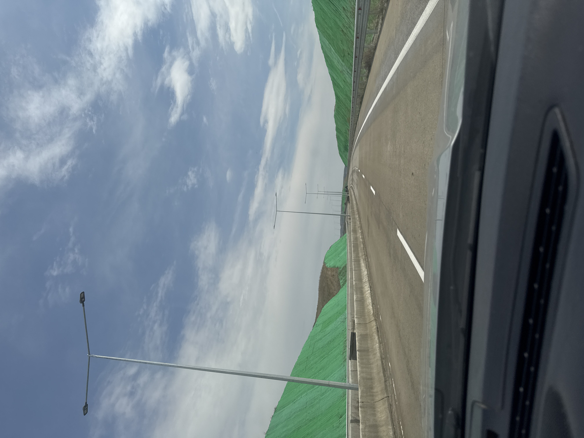 The image shows a view from inside a moving vehicle, likely on a highway. The road stretches into the distance with white lane markings and a shoulder line. Hills covered in green vegetation flank the road on the right. Streetlights are visible along the highway. The sky is partly cloudy. The location is Mukhrovani, Georgia.