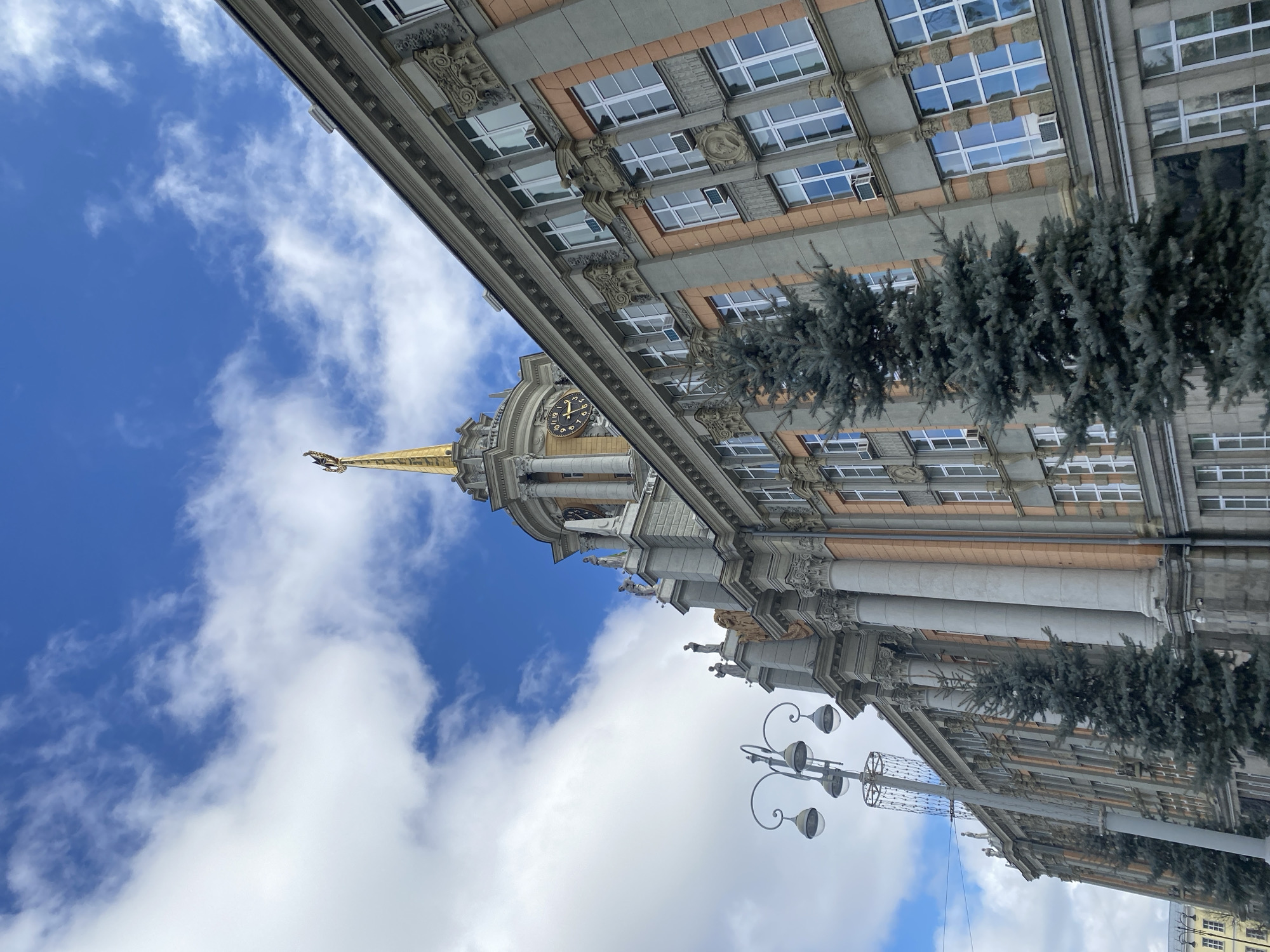 The image captures a view of the City Administration Building in Yekaterinburg, Russia, tilted with the building dominating the right half of the frame. The architectural style is grandiose, featuring ornate details, columns, statues, and rows of windows. A clock tower with a gold spire extends upwards towards a partly cloudy, blue sky. Rows of pine trees are visible along the building's façade. A street lamp is visible in the lower-left portion of the frame. The sky is a mix of blue and white clouds.
