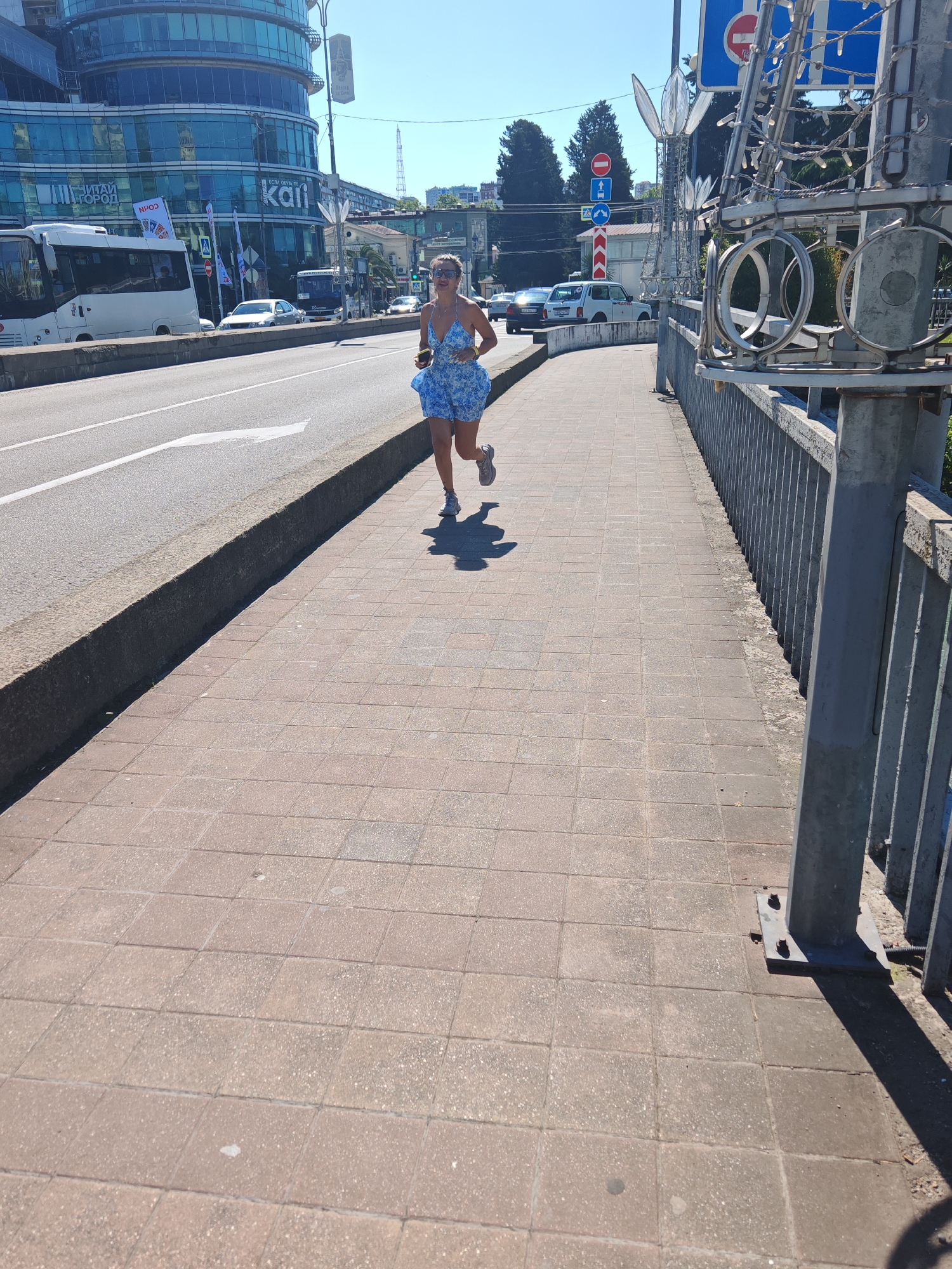 An outdoor, eye-level shot captures a woman running on a paved sidewalk next to a busy urban road on a bright, sunny day. The woman, wearing a blue and white patterned mini-dress and running shoes, is in motion, with sunglasses pushed up onto her head and what appears to be a yellow device in her hand. Her shadow is cast long on the reddish-brown square-tiled sidewalk, indicating a high sun.
To her left, a multi-lane asphalt road is visible, with a white arrow painted on the surface and a concrete barrier separating it from the sidewalk. Several vehicles, including a white bus and cars, are on the road in the background.
The background features a large, modern building with a curved glass facade reflecting the sky, displaying the partial word "kali" and banners with Cyrillic text, including "ЧИТАЙ ГОРОД" (CHITAI GOROD) and "НПОД", as well as "ЕСЛИ ОБУЧ". Further down the road, more buildings and trees are visible.
To the right, a grey metal railing runs alongside the sidewalk, adorned with decorative metallic structures, possibly for lighting. Traffic signs, including "No Entry" and "Turn Right" signs, are visible on the far right. The clear blue sky and strong shadows suggest it is midday or early afternoon. The overall scene depicts a bustling city environment.
