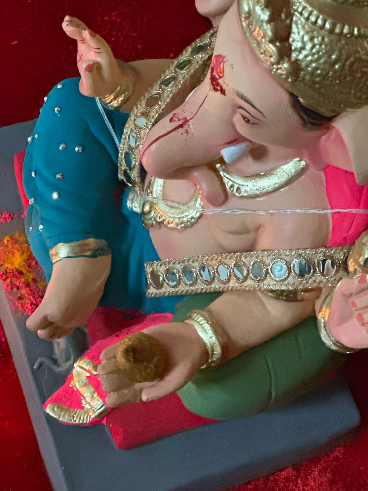 This image shows a close-up, eye-level shot of a statue of the Hindu deity Ganesha, likely located in Chhatrapati Sambhajinagar, India. The statue is ornately decorated, with a golden headdress and jewelry. Ganesha's trunk is turned to the right, and he is holding a small, round object in his right hand. The statue is wearing a blue and pink garment adorned with small, shiny embellishments and a golden, mirror-studded belt. The background is a plain red surface. The lighting is artificial and appears to be coming from above, creating soft shadows. The overall impression is one of vibrant color and detailed craftsmanship.