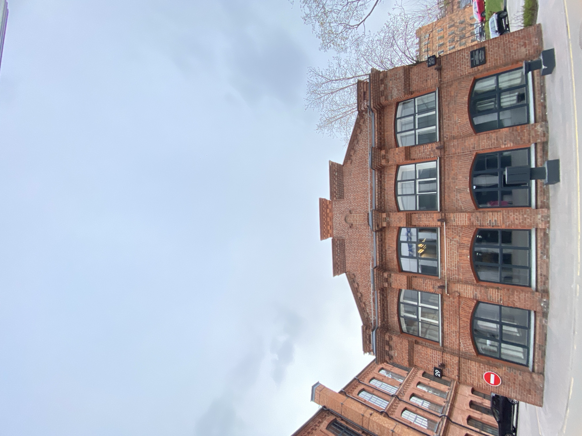The image shows a low-angle view of a red brick building against a cloudy sky. The building has arched windows, and there is a "no entry" sign visible on the lower right corner. The building appears to be in Moscow, Russia, specifically the Trekhgornaya Manufactura historical industrial complex.