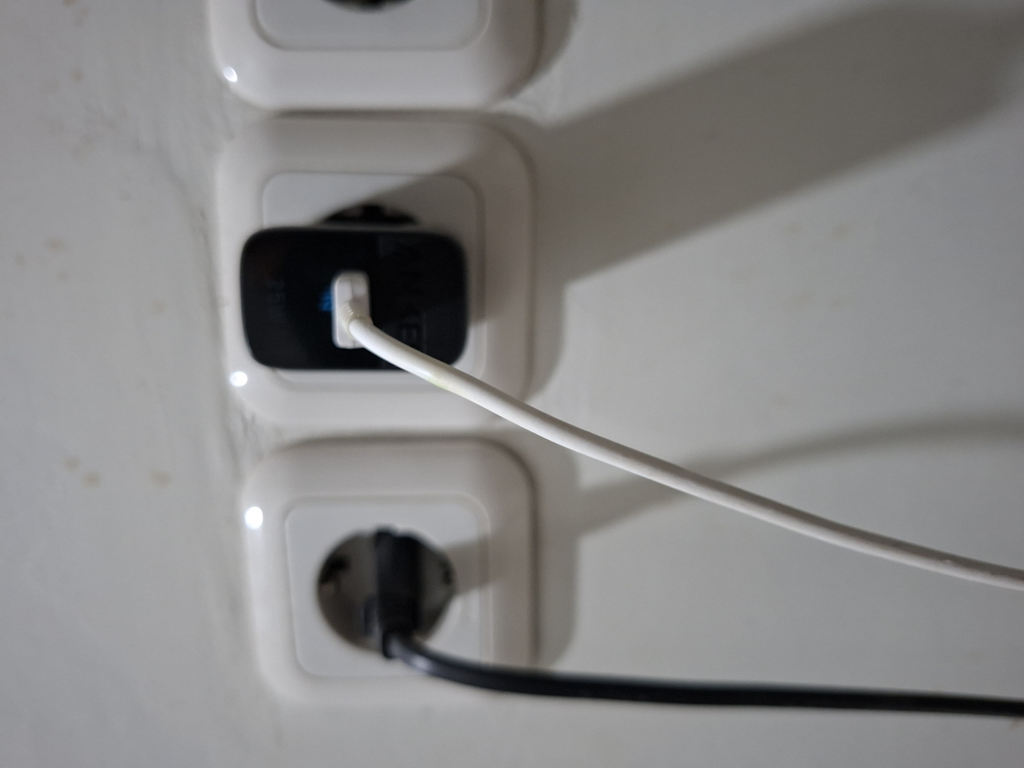 The image shows a close-up of three electrical outlets on a white wall. A black electrical cord is plugged into the leftmost outlet. A white cable is plugged into the middle outlet, connected to a black charging adapter. The rightmost outlet is empty. The wall appears to have some minor discoloration or stains. The lighting suggests an indoor setting. The image is taken from a slightly elevated angle. There is no indication of any specific location in Samarinda, Indonesia, as the scene is generic and lacks identifying features.