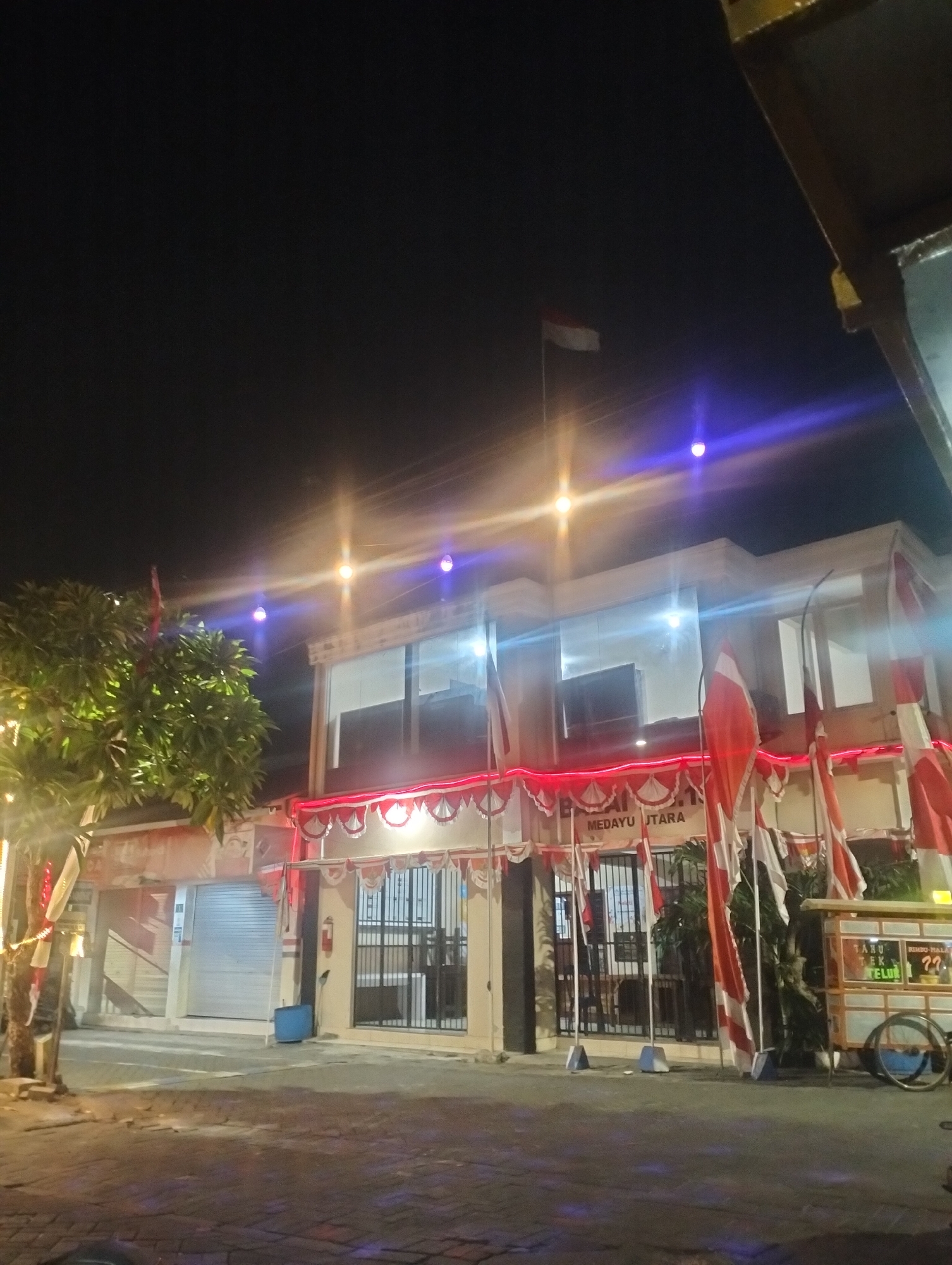 The image depicts a nighttime scene in front of a building adorned with Indonesian flags and red and white decorative bunting. The building appears to be a commercial establishment or government office, with the text "MEDAYU UTARA" visible on its facade. In front of the building, there are several flags, some of which are the Indonesian flag, red and white. There is a tree on the left side of the image, illuminated by lights, and a food cart with wheels on the right. The overall impression is festive, possibly in celebration of a national holiday. The location is identified as Medayu Utara, a subdistrict in Surabaya, Indonesia.