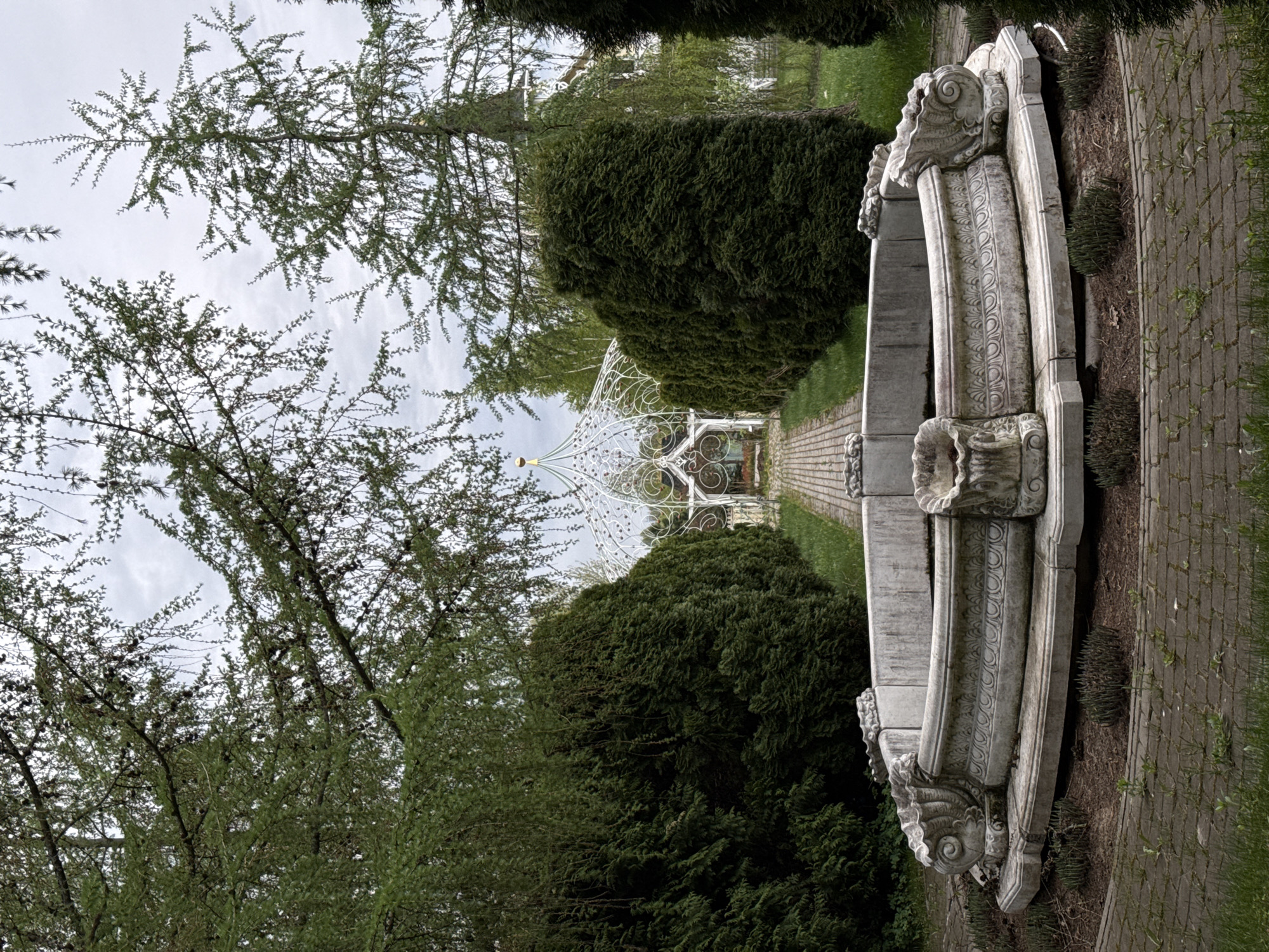 The image shows a park scene in Sapronovo, Russia, featuring a stone fountain in the foreground. Behind the fountain, a pathway leads towards a white, ornate gazebo. Green, rounded bushes line the pathway, flanked by trees. The sky is overcast.