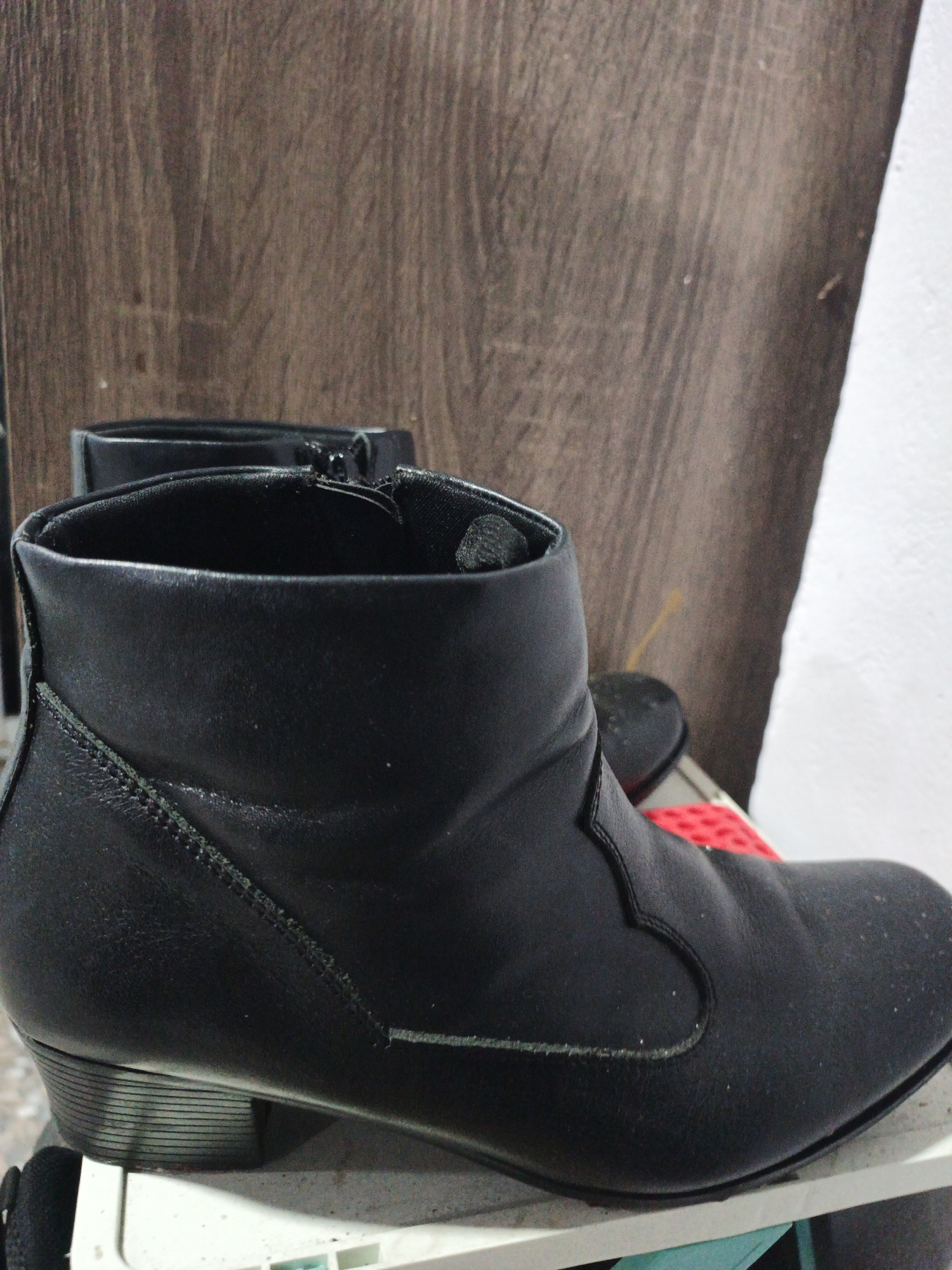 A pair of black leather ankle boots with a side zipper are the primary focus of the image. The boots are positioned on a white shelf in front of a wooden-patterned background. The lighting is dim, and the overall scene appears to be indoors, likely in a residential or storage area. The boots exhibit some dust accumulation, suggesting they are not in active use or have been stored for some time. There is no indication of any specific location within Kelapapati, Indonesia, beyond the general indoor setting. The image does not contain any people, actions, or interactions.