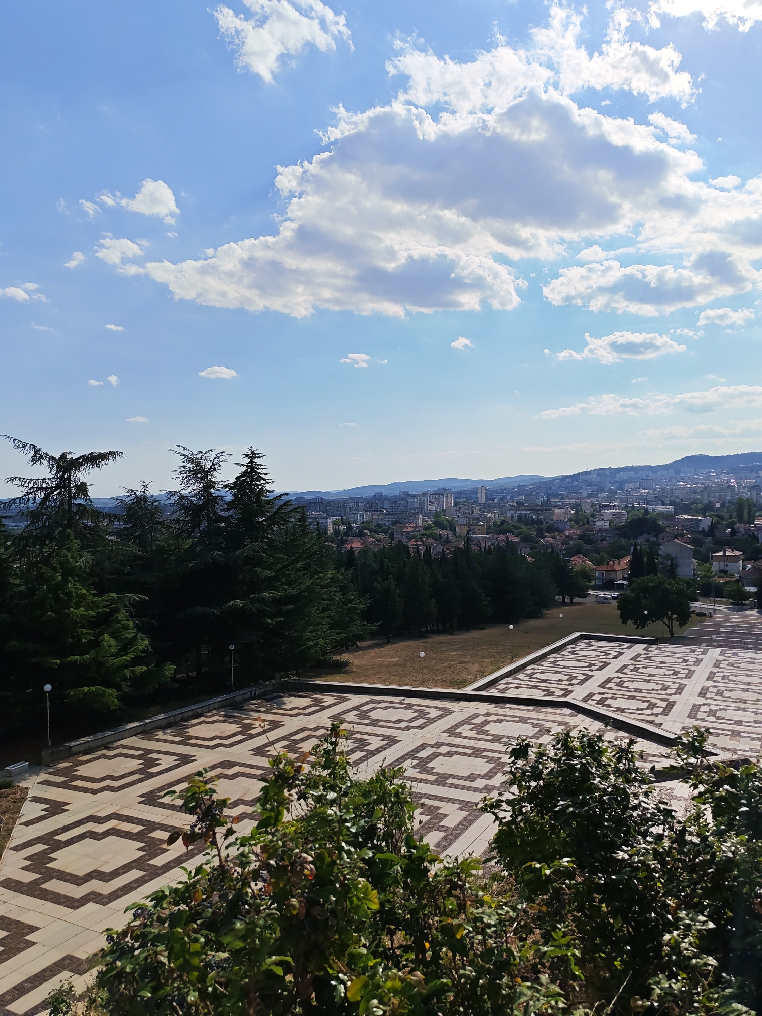 The image captures a panoramic view from a high vantage point, overlooking the city of Stara Zagora, Bulgaria. In the foreground, a large, patterned paved area with geometric designs is visible, bordered by lush green foliage. Behind this, a dense cluster of tall evergreen trees, likely cypress or fir, forms a dark green barrier. Beyond the trees, the city unfolds with a mix of buildings, ranging from residential structures with red-tiled roofs to taller, modern apartment buildings and other urban constructions. The cityscape extends to the horizon, where rolling hills under a bright blue sky dotted with fluffy white clouds are seen. The overall scene suggests a clear, sunny day. The specific location is likely the area around the "Osnova" monument, which features a similar patterned plaza and overlooks the city from a prominent hill.
