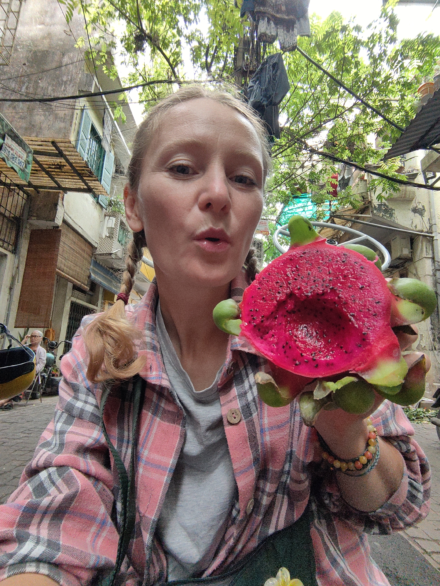 A cheerful young woman takes a close-up selfie, prominently holding a vibrant, partially eaten dragon fruit in an urban alleyway in Hà Nội, Vietnam. She has blonde hair styled in two pigtail braids and wears a pink, grey, and white plaid shirt over a grey t-shirt. Looking directly at the camera, her mouth is slightly open, as if blowing a kiss or expressing delight. Her right wrist is adorned with several colorful beaded bracelets.
The dragon fruit is striking, with its bright pink skin and green scales, revealing a vivid dark pink interior speckled with tiny black seeds, where a portion has been scooped out.
The background shows a narrow, bustling street or alley with multi-story buildings featuring balconies and traditional shutters. Lush green tree foliage overhead filters the daylight, and numerous overhead wires crisscross the scene. In the distance on the left, an elderly person with white hair is faintly visible sitting down. A green sign in the upper left displays a partial phone number "916.083.967" and the beginning of a Vietnamese word, "DU LỊCH..." (Tourism). The bright natural light suggests it is daytime.
