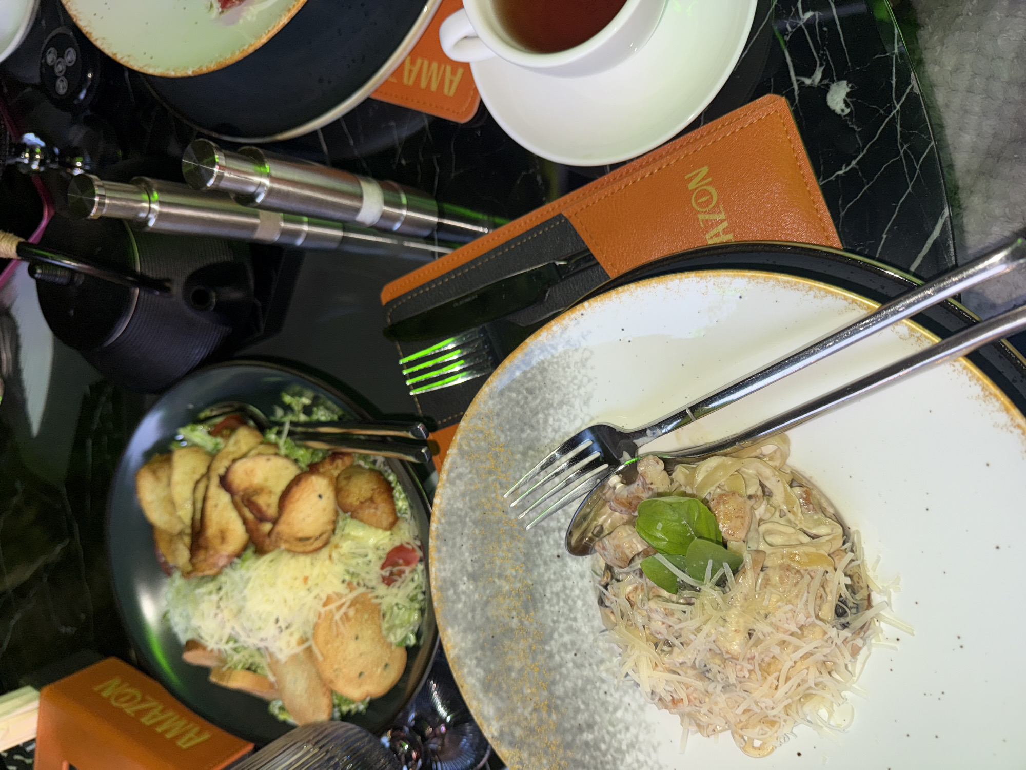 The image captures a dining setting, likely in a restaurant or cafe in Yerevan, Armenia. Two plates are visible: one holds a salad with fried croutons and shredded cheese, while the other contains pasta topped with cheese and basil leaves. A cup of tea rests on a coaster. Silverware is placed on the pasta dish. The word "AMAZON" is visible on the coaster and another item, suggesting branding. Light reflects off shiny metal objects and a dark, patterned table.