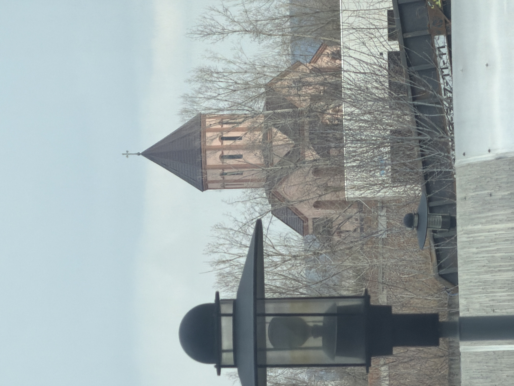 A low-angle shot depicts a section of the Church of the Intercession in Krasnoyarsk, Russia. The church features a tall, multi-story tower with a brown roof topped by a cross. The building's walls are light orange with vertical red stripes at each corner. A streetlight occupies the lower foreground, with its black lamp post and enclosed bulb. Bare tree branches partially obscure the view of the church. A white wall and a dark, vertical structure are partially visible on the right. The sky is overcast.