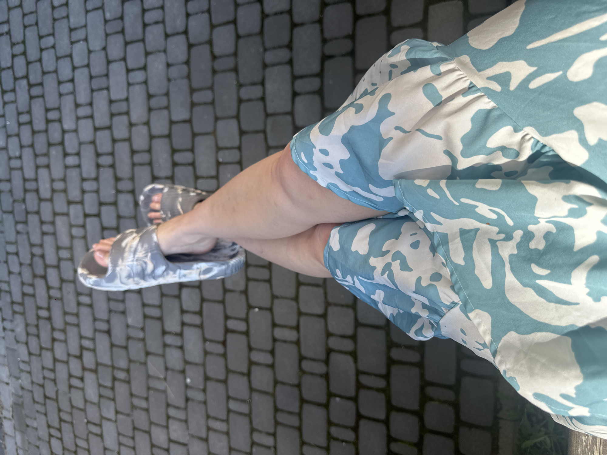 A person is sitting outside with their legs crossed and resting on a wooden surface. They are wearing a blue and white patterned dress and grey and white patterned slides. The background consists of a dark grey brick patio. The lighting suggests it is daytime.