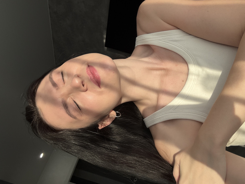 A young woman with dark hair and closed eyes lies reclined in a casual white tank top. Sunlight casts shadows across her face. She has fair skin, subtle pink lipstick, and wears a delicate silver hoop earring. Her hand rests near her chest. The background features a dark, glossy surface and a light gray wall, adding contrast to the subject. The picture is composed with a tilted angle, creating a relaxed, informal feel.