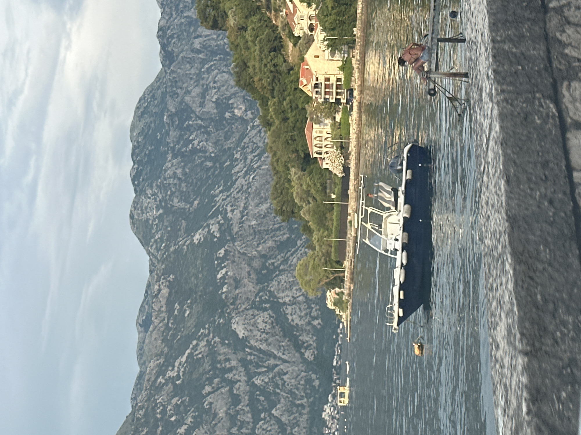 In Prčanj, Montenegro, on a body of water with mountains in the background, a blue and white motorboat is docked. To the right, a person sits on a wooden structure over the water. In the distance, houses with red roofs are visible along a treed shoreline, with a yellow floating structure further out on the water. The sky is cloudy.