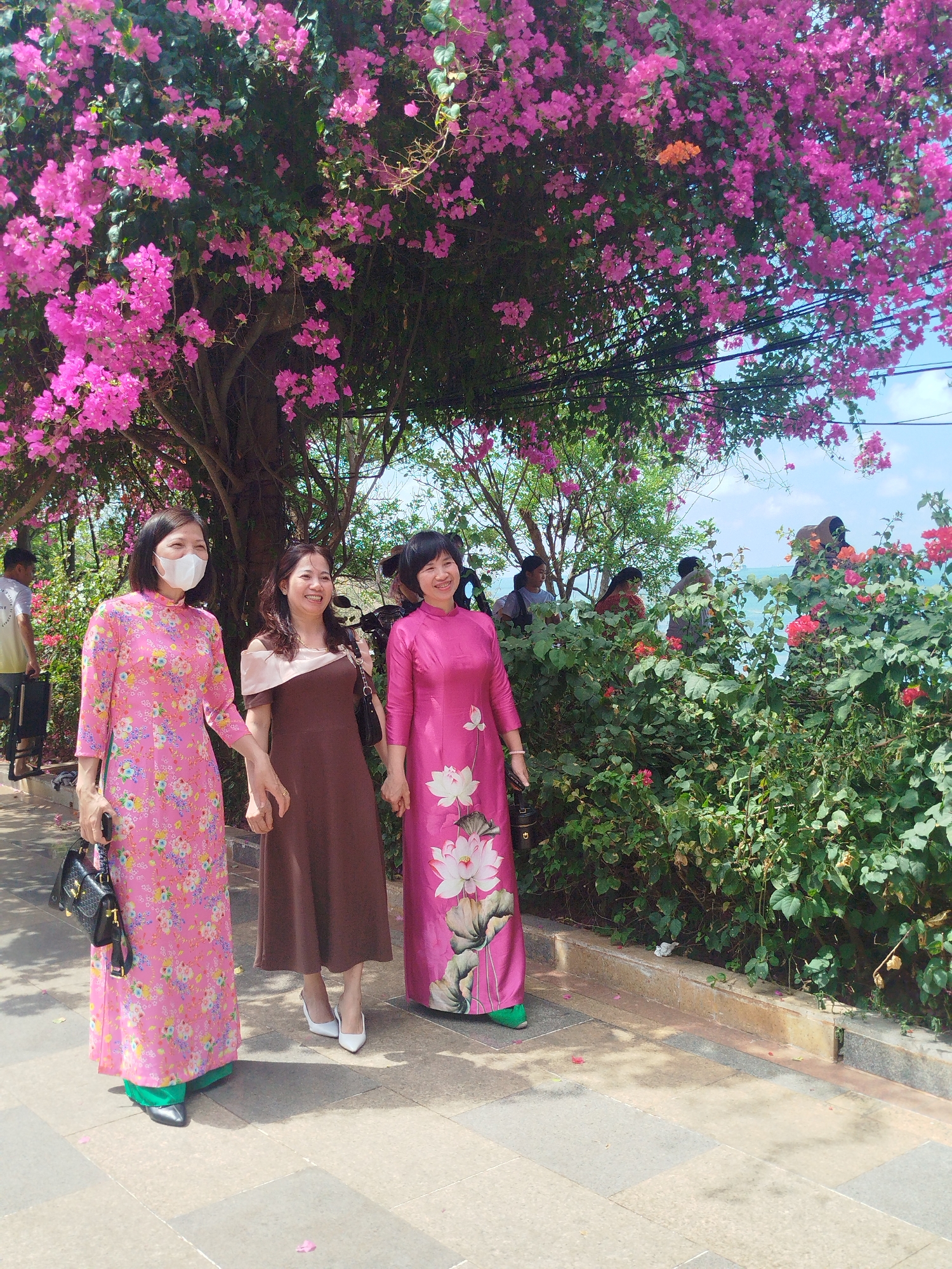 Three women are walking together along a sidewalk near a body of water, likely the ocean, in what appears to be a public park or garden. The woman on the left is wearing a pink floral patterned dress with long sleeves, a white face mask, and carrying a black purse. The middle woman is wearing a brown dress with off-the-shoulder detail and white high heels. The woman on the right is wearing a bright pink long-sleeved dress with a large white floral design on the front. They are holding hands and smiling.
Above them is a large tree with vibrant pink flowers. To the right of the women is a large bush with red flowers. In the background, there are other people and some water with boats in the distance. The scene is well-lit with natural daylight. The ground is paved with light-colored stones or tiles. The location is likely a coastal area or park within Ho Chi Minh City, Vietnam.