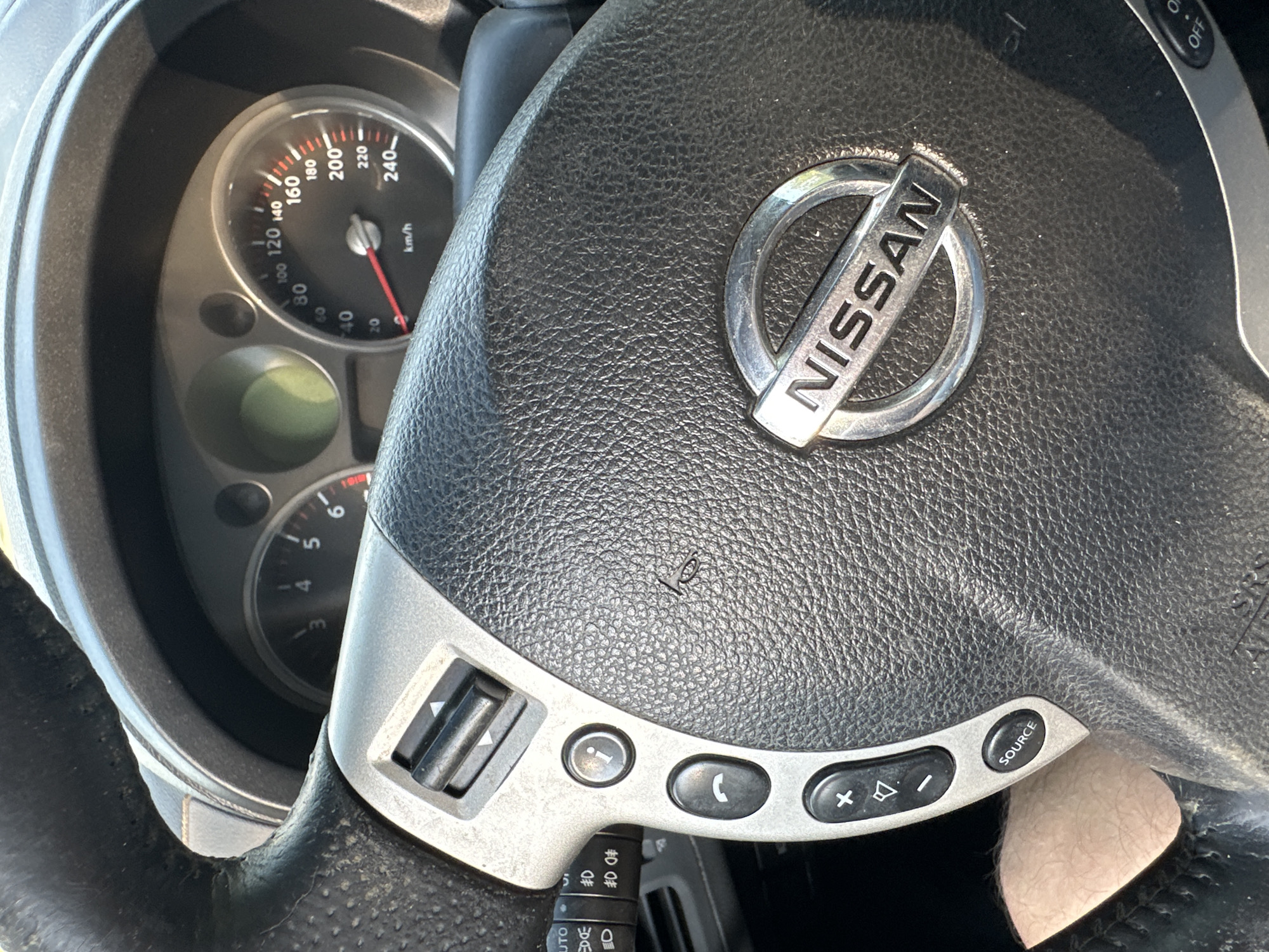 The image shows a close-up of a car's steering wheel and dashboard. The steering wheel is black leather with silver accents and has several buttons on it, including a volume control, a call button, and a "SOURCE" button. The Nissan logo is prominently displayed in the center of the steering wheel. The dashboard includes a speedometer that reads approximately 40 km/h. The car appears to be stationary. There is no indication of the location being Shatava, Ukraine.
