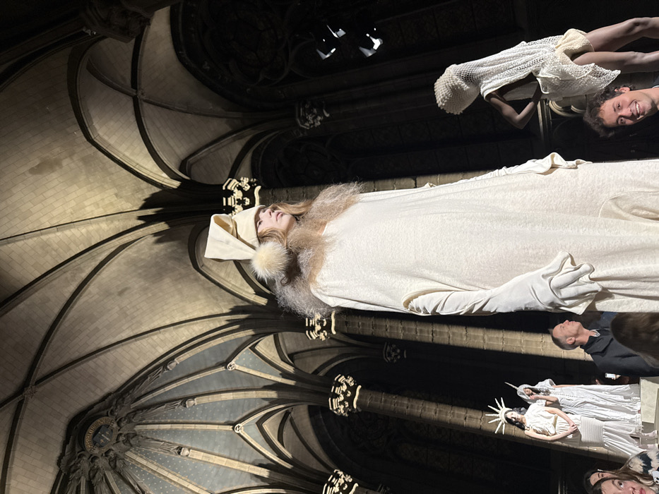 A fashion show unfolds inside a gothic cathedral. A model in a long white dress with gloves and furry collar is center stage. Other models wear white clothing with quirky headwear resembling crowns or the Statue of Liberty. Guests are seen watching the show. The architecture features pointed arches and ribbed vaults, lit to accentuate the space. Two spotlights shine from the ceiling.