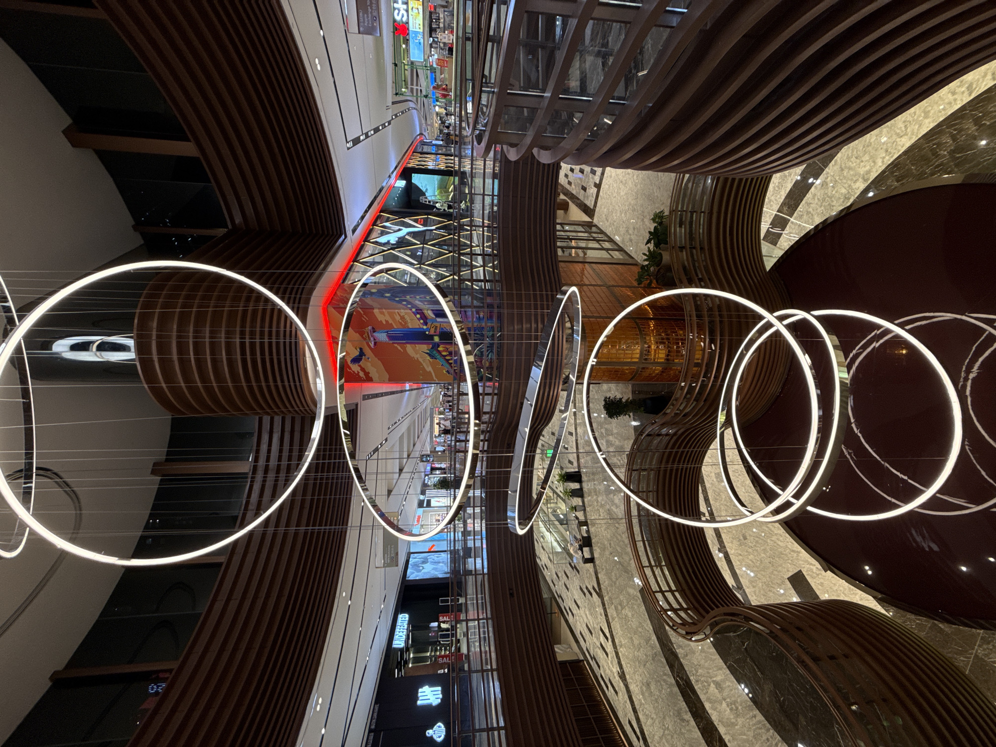 The image depicts an interior view of a modern shopping mall, likely in Kuala Lumpur, Malaysia, characterized by a multi-story atrium with contemporary architectural elements. The focus is on a series of large, circular LED light fixtures suspended from the ceiling, creating an artistic and ambient lighting effect.
The mall's interior features a blend of materials, including wooden slat structures, glass panels, and marble flooring. Various shops and retail spaces are visible along the upper levels, indicated by storefront signs and displays. People are observable in the distance, suggesting normal pedestrian traffic within the mall. A combination of natural and artificial light sources illuminates the space. The composition and perspective suggest that the photograph was taken from an elevated vantage point.