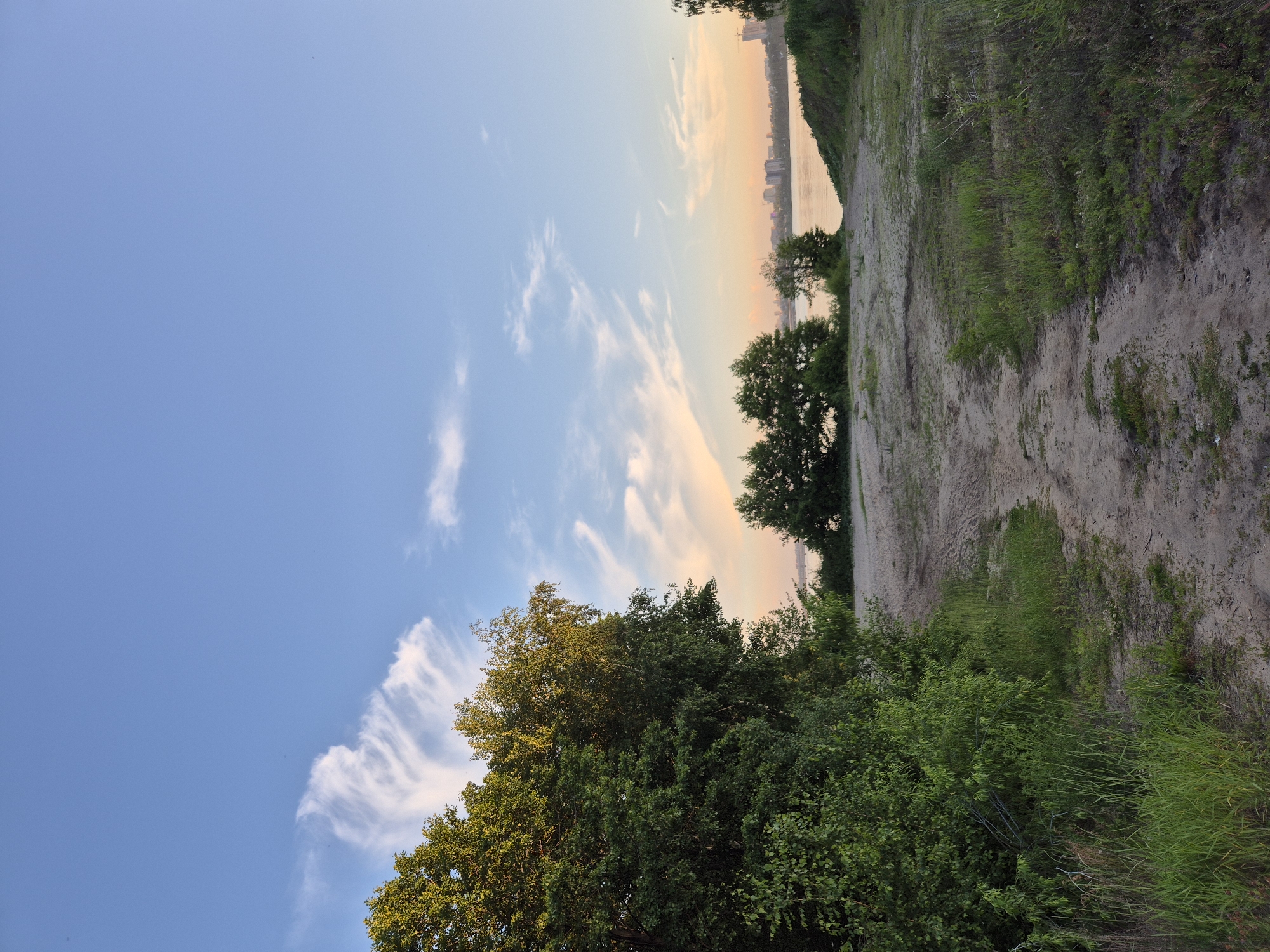 An outdoor, low-angle, medium shot shows a sandy path leading towards a body of water at dusk. The sky is a light blue with wispy white and pink clouds. On the left, dense green foliage and trees border the path. In the distance, across the water, a city skyline is visible with buildings and structures. The path is made of light-colored sand with patches of grass and some tire tracks. The overall mood is peaceful and serene.