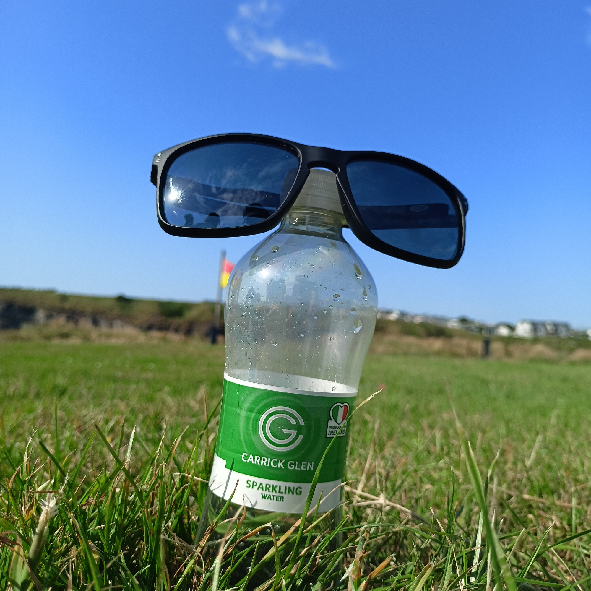 A close-up, low-angle shot captures a partially filled, clear plastic bottle of Carrick Glen Sparkling Water in the foreground, nestled in green grass under a bright blue sky. The bottle has a green label with white text and a circular 'G' logo, as well as a small heart graphic with the Italian flag and the word "PRODUCED" and "IRELAND" below it. The neck of the bottle is topped with a white cap. Perched on top of the bottle are a pair of dark-rimmed sunglasses, with their lenses reflecting the sky and some indistinct shapes, possibly including a silhouette of the photographer.
In the mid-ground, a slightly out-of-focus landscape unfolds. A gently sloping grassy hill rises behind the bottle, and on top of it, a red and yellow flag is visible on a pole. Further in the distance, a line of houses or buildings can be seen along the crest of the hill. The overall scene suggests an outdoor setting, likely a park or open field, on a sunny day. The location is identified as Ballybunion, Ireland.