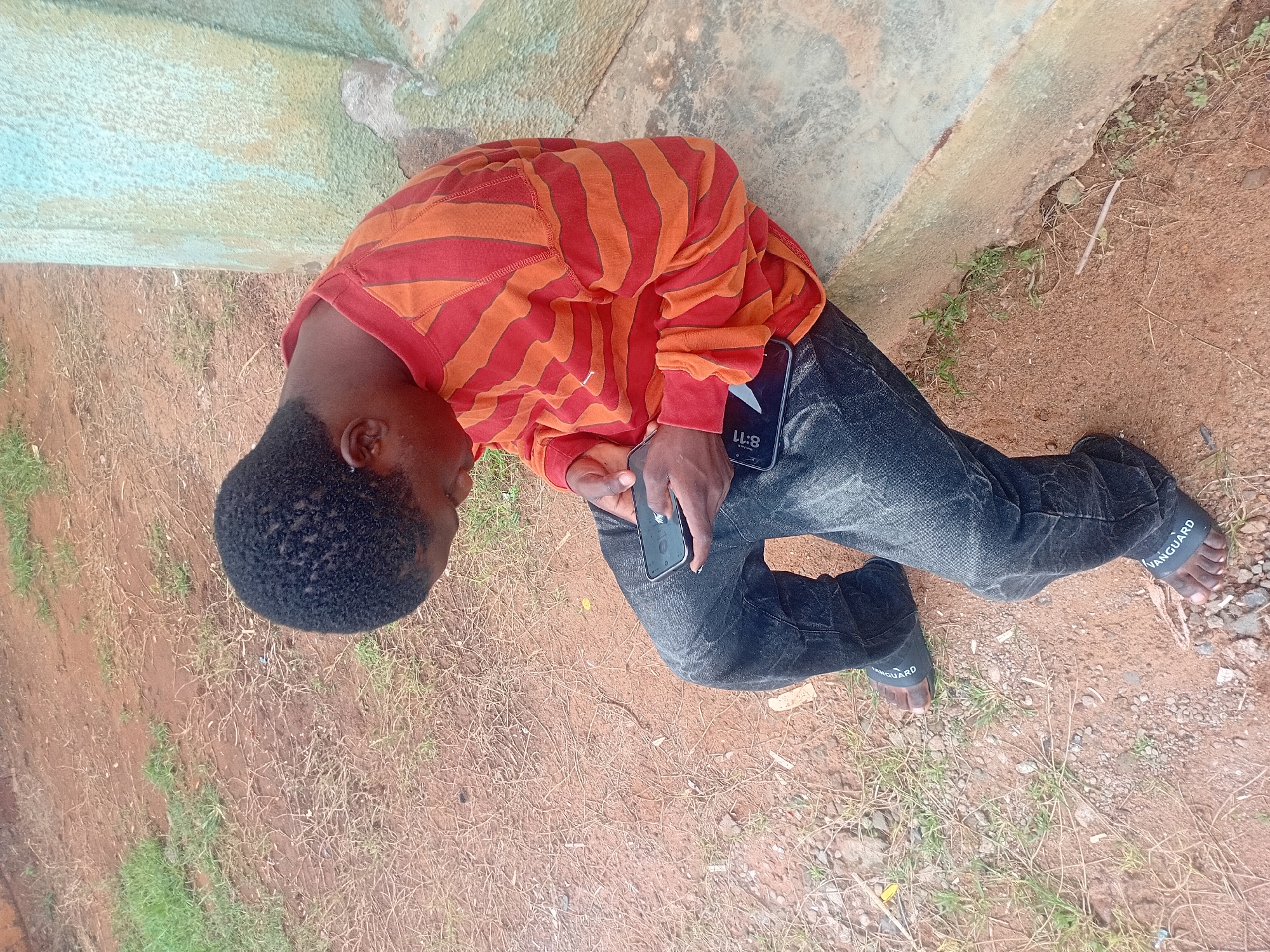 A young Black male wearing a red and orange striped long-sleeved shirt and dark wash jeans sits on a concrete ledge. He is looking down at a smartphone in his hands. His feet are bare, except for black sandals with the word "VANGUARD" printed on them. The ground around him is a mixture of dry dirt, small rocks, and sparse dry grass. To his right, a light blue and green painted concrete structure rises vertically. The scene appears to be outdoors in a non-urban area, possibly a backyard or undeveloped plot in Ogbogbo, Nigeria, indicated by the red soil, sparse vegetation, and the style of the concrete structure.