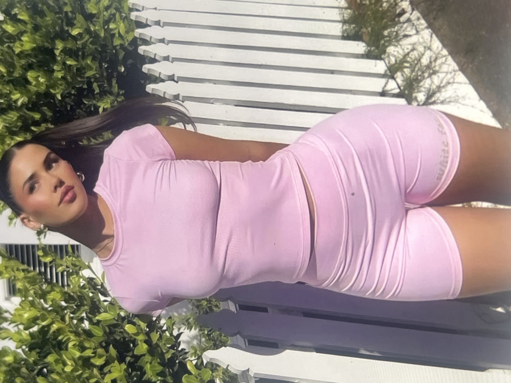 A woman wearing a pink t-shirt and pink shorts stands outside in front of a white picket fence. Green foliage is visible behind her and to her left. The woman has long dark hair pulled back into a ponytail and is wearing hoop earrings. The lighting suggests it is daytime.