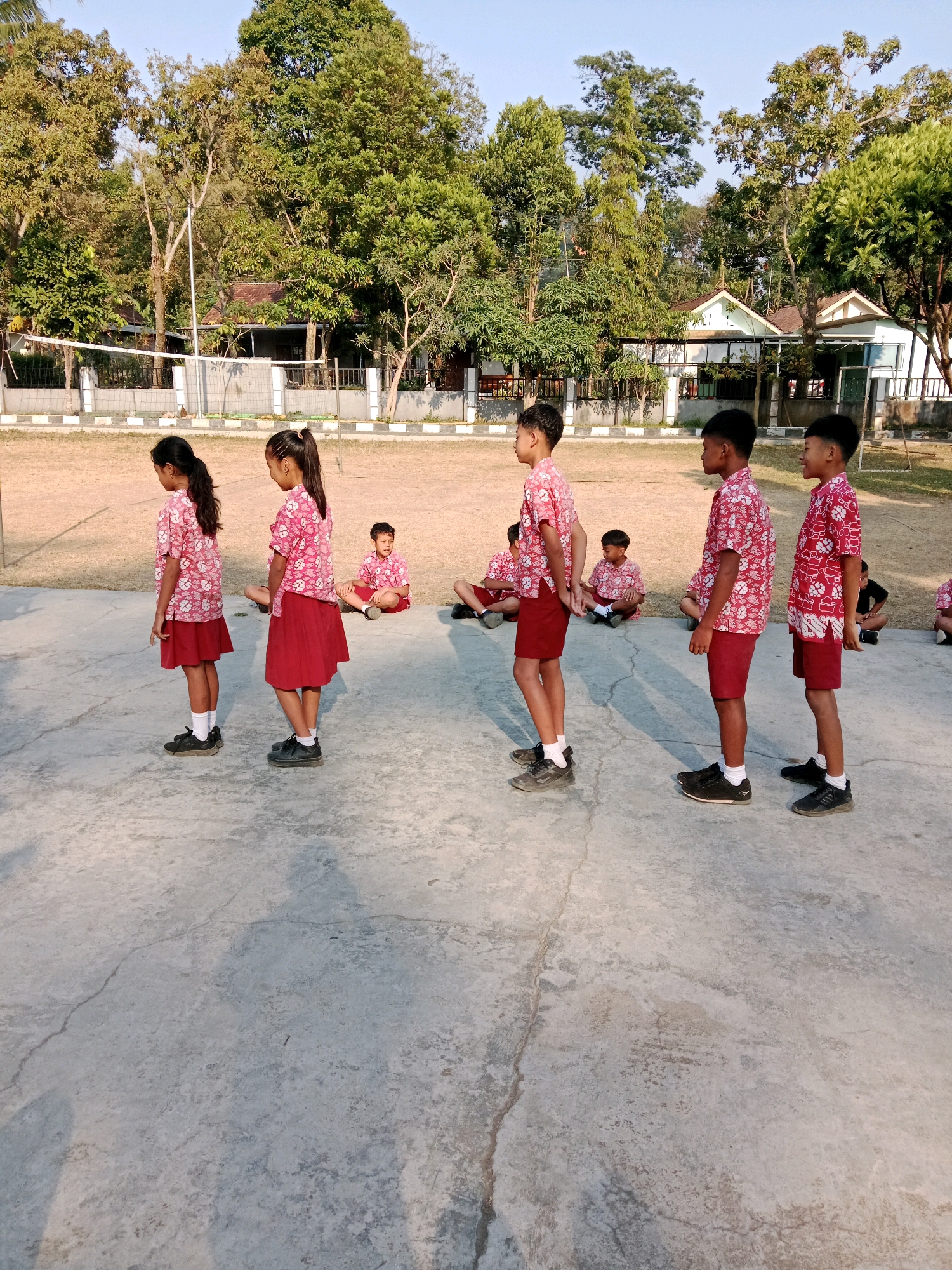 In Salatiga, Indonesia, a group of children in uniform are engaged in an outdoor activity on a concrete surface. Six children stand in a line, facing right. The first three children from the left are girls, wearing pink batik shirts and red skirts. The next three children are boys, also in pink batik shirts and red shorts. Behind them, several other children are seated on the ground. In the background, a volleyball net and court are visible, along with trees and buildings, suggesting a school or community area. The lighting indicates it is daytime, likely morning or afternoon, with shadows cast on the ground.