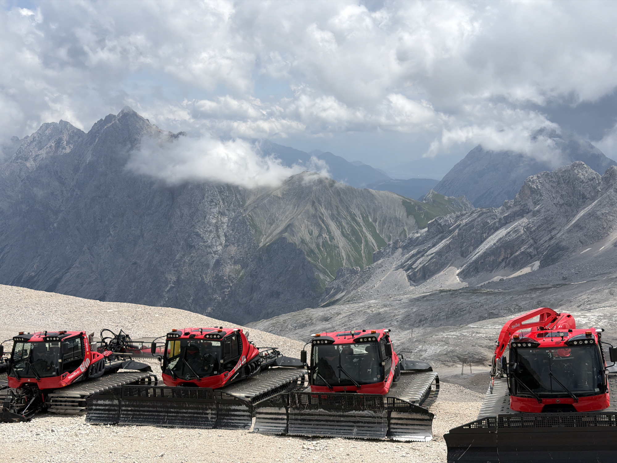 In Garmisch-Partenkirchen, Germany, on a mountain summit, four bright red snow groomers are parked on a rocky, gravelly terrain. The machines, with their articulated tracks and large cabins, are positioned in a line, facing towards the viewer and slightly to the right. The foremost snow groomer on the right has a red robotic arm extended upwards. In the background, rugged mountain peaks rise, some partially obscured by dramatic clouds. The sky is a mix of grey and white clouds, with patches of blue visible. The scene suggests a high-altitude, possibly summer, environment where snow groomers are maintained or prepared for winter use.
