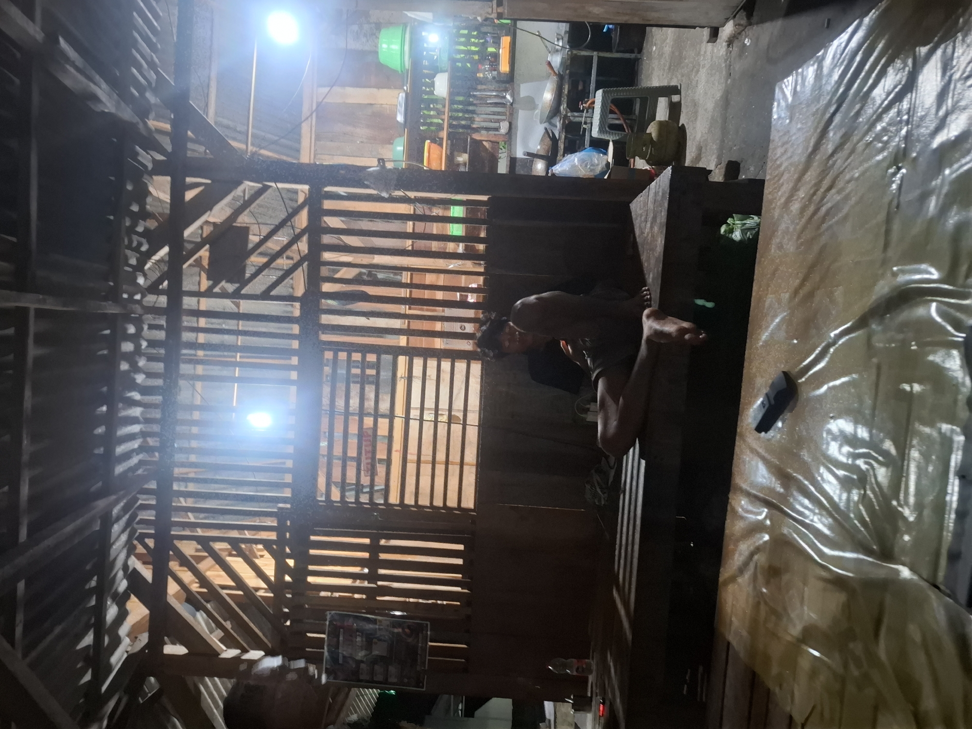The image depicts a dimly lit interior space, likely a semi-open structure with a corrugated metal roof and wooden plank walls and ceiling. The setting appears to be a modest dwelling or workshop in Bulubrangsi, Indonesia.
In the center of the frame, a person is seated on a low wooden platform, facing left. They are wearing dark clothing and appear to be holding a mobile phone. Their legs are extended, with one foot resting on the platform. The person's posture suggests relaxation or casual activity.
To the left of the seated person, on the same platform, is a clear plastic bottle with a white cap. Further left, a poster with text and images is affixed to the wall.
In the background, to the right of the seated person, a shelf unit is visible, stocked with various items including green bowls, plastic containers, and bottles. A gas cylinder and a small stool are also present in the background, suggesting a functional area, possibly a kitchen or storage space. The lighting in the scene comes from overhead bulbs, casting long shadows across the wooden floor and platform. The overall impression is one of a utilitarian and lived-in environment.